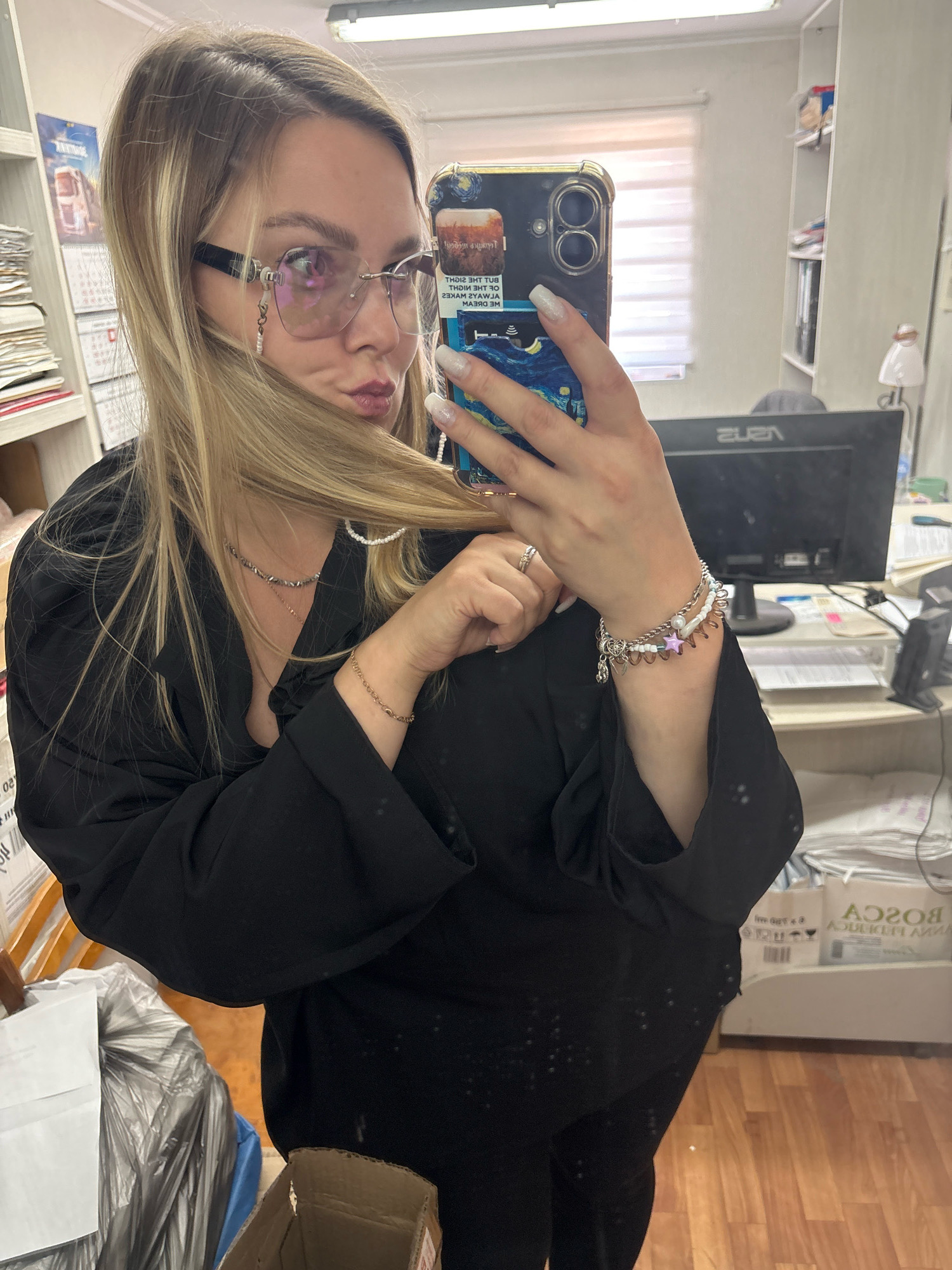 A woman with blonde hair and light-colored sunglasses is taking a selfie in an office environment. She is wearing a black top and has multiple bracelets on her left wrist. The background includes shelves with papers and office supplies, a computer monitor, and a trash can. The location is identified as Noviy, Russia.