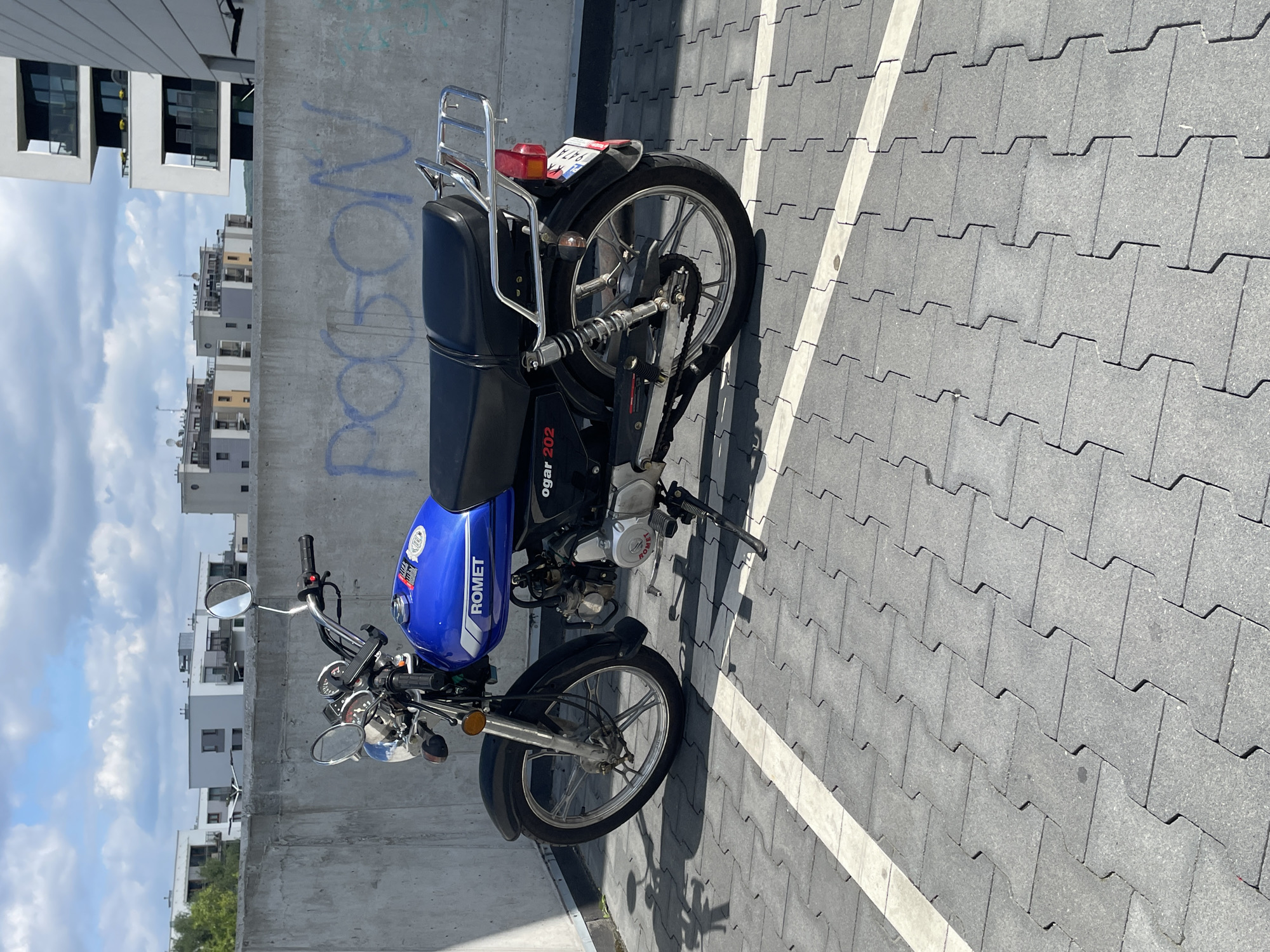 A blue Romet Ogar 202 motorcycle is parked on a gray interlocking paver surface. The motorcycle is positioned at an angle, facing towards the right side of the frame. It has a black seat and a chrome luggage rack at the rear. The motorcycle's license plate is visible. Behind the motorcycle, a concrete wall with graffiti is present. In the background, there are modern buildings and a cloudy blue sky. The scene appears to be an outdoor parking area.
