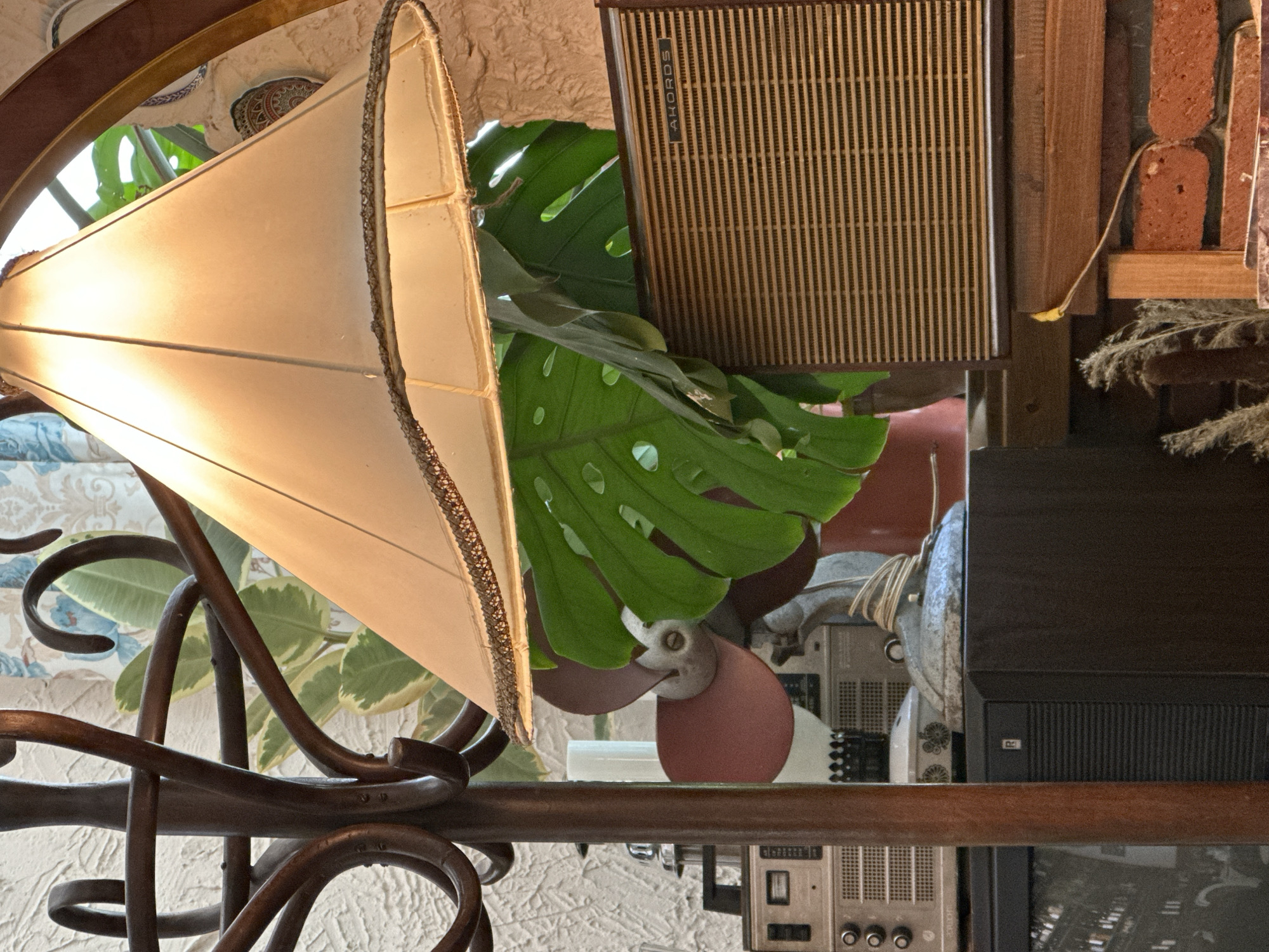 A cluttered interior scene with a variety of objects. Dominating the left is a lit, tan lampshade with a decorative rim, attached to a brown wrought-iron frame. Behind the lampshade, a large green monstera leaf adds a botanical element. To the right of the leaf is a square, light brown speaker with vertical dark stripes. Beneath the leaf and speaker, a vintage fan with red blades and a metallic casing is partially visible. Below the fan and to the right, several electronic devices, possibly radios or similar equipment, are stacked, with a black television screen partially visible at the bottom. In the background, a textured wall with a decorative pattern can be seen, and glimpses of additional objects like a brick wall, wooden shelf, and various plants suggest the scene may be in an eclectic Tbilisi, Georgia cafe or antique shop. The scene is lit with warm tones, creating a cozy atmosphere.