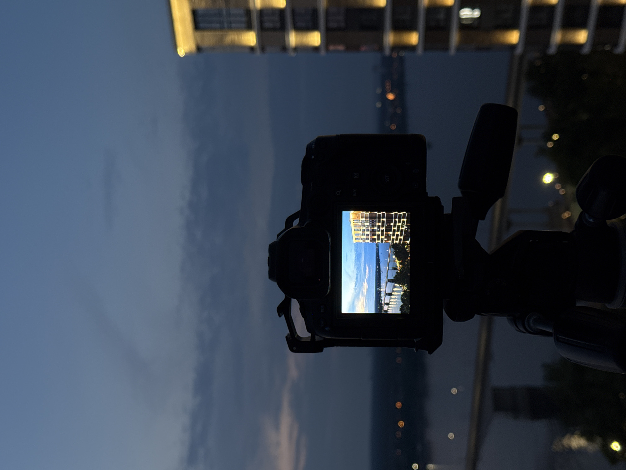 The image shows a camera on a tripod, taking a photo of a city skyline at dusk. The camera's screen displays a brightly lit modern building and a bridge spanning a body of water. In the background, the sky is a deep blue, with hints of clouds. The body of water reflects the lights of the city. The building in the foreground, on the right side of the image, is tall and modern, with lights illuminating its facade. This is likely an evening shot captured from a vantage point overlooking the Kama River in Perm, Russia, given the architectural style of the buildings and the presence of a prominent bridge.