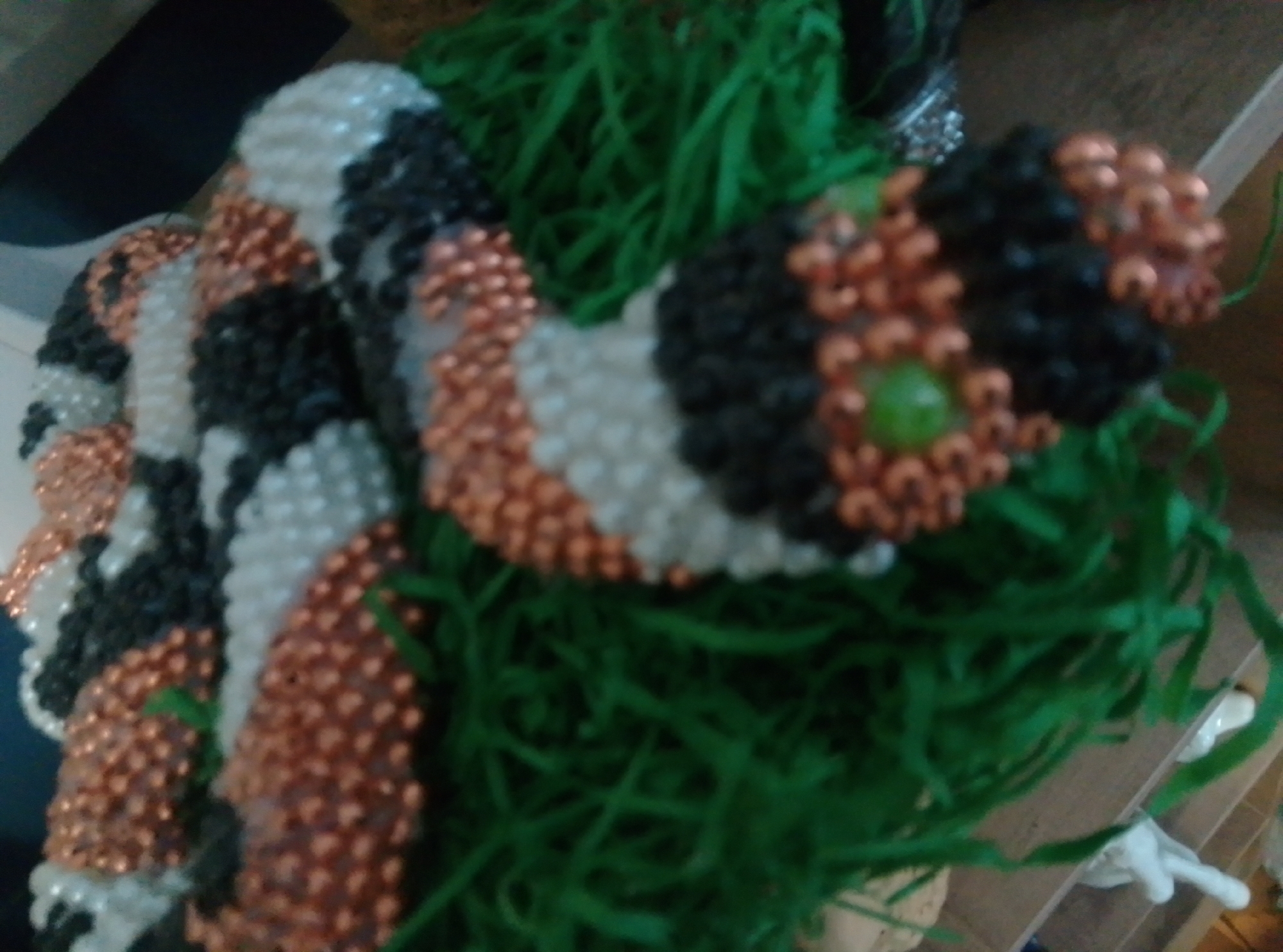 The image shows a beaded object, possibly a craft or decoration, resembling a coiled snake. The object is predominantly black, white, and copper-colored, with some green bead accents. It is situated amidst a bed of artificial green grass. The background is blurred, but appears to be an indoor setting with wooden surfaces. There are no people or discernible actions visible in the image. The image does not contain information about the city of Wedel, Germany.
