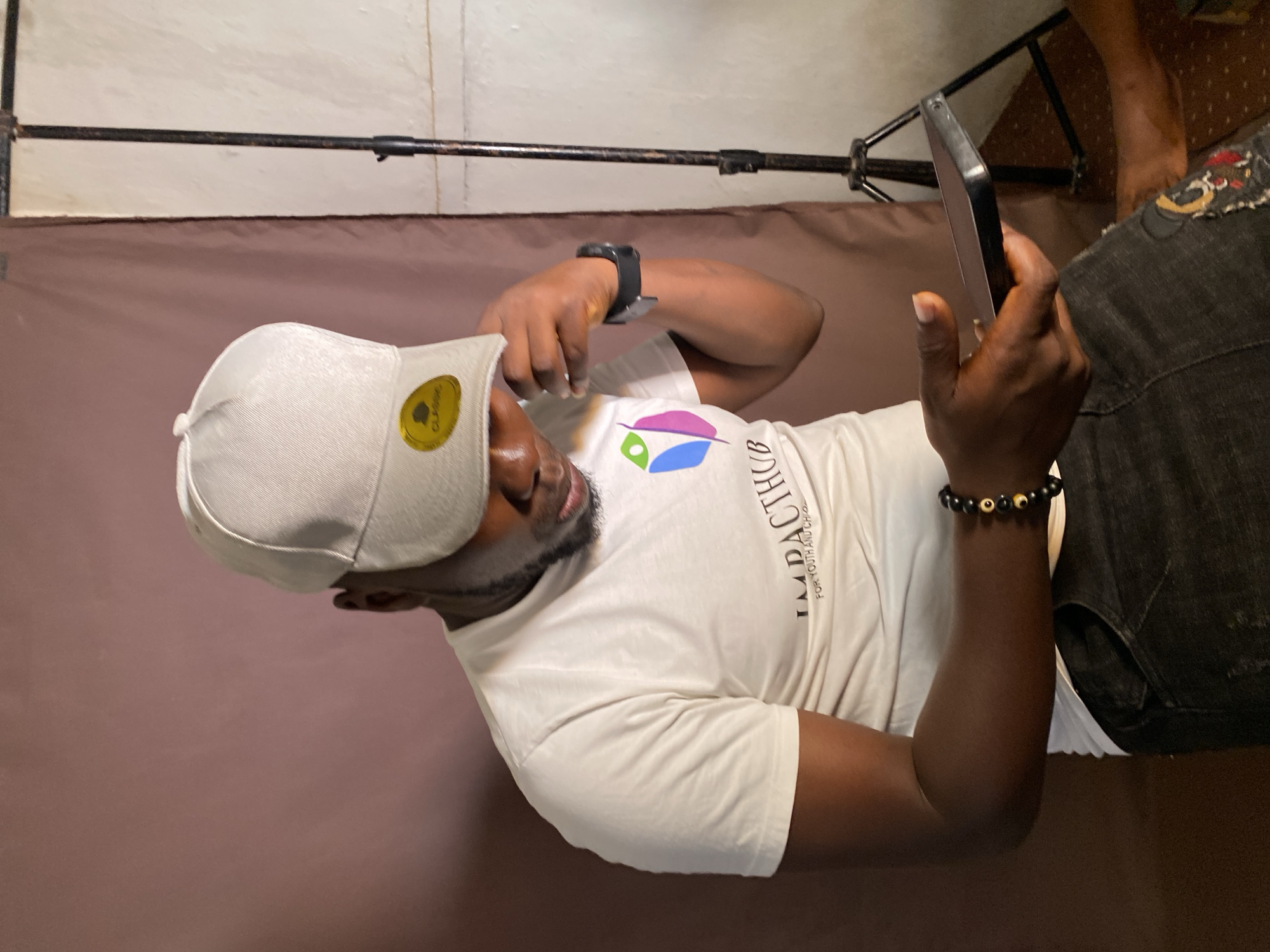 A man, wearing a white baseball cap, a white t-shirt with "IMPACTHUB FOR YOUTH AND CHILDR" printed on it, a black watch, and a bracelet with dark beads and light-colored accents, is looking at a black smartphone held in his right hand. He is lying on a brown backdrop. A black metal bar is above him. He is also wearing dark jeans with a decorative patch. The location is Vom, Nigeria.