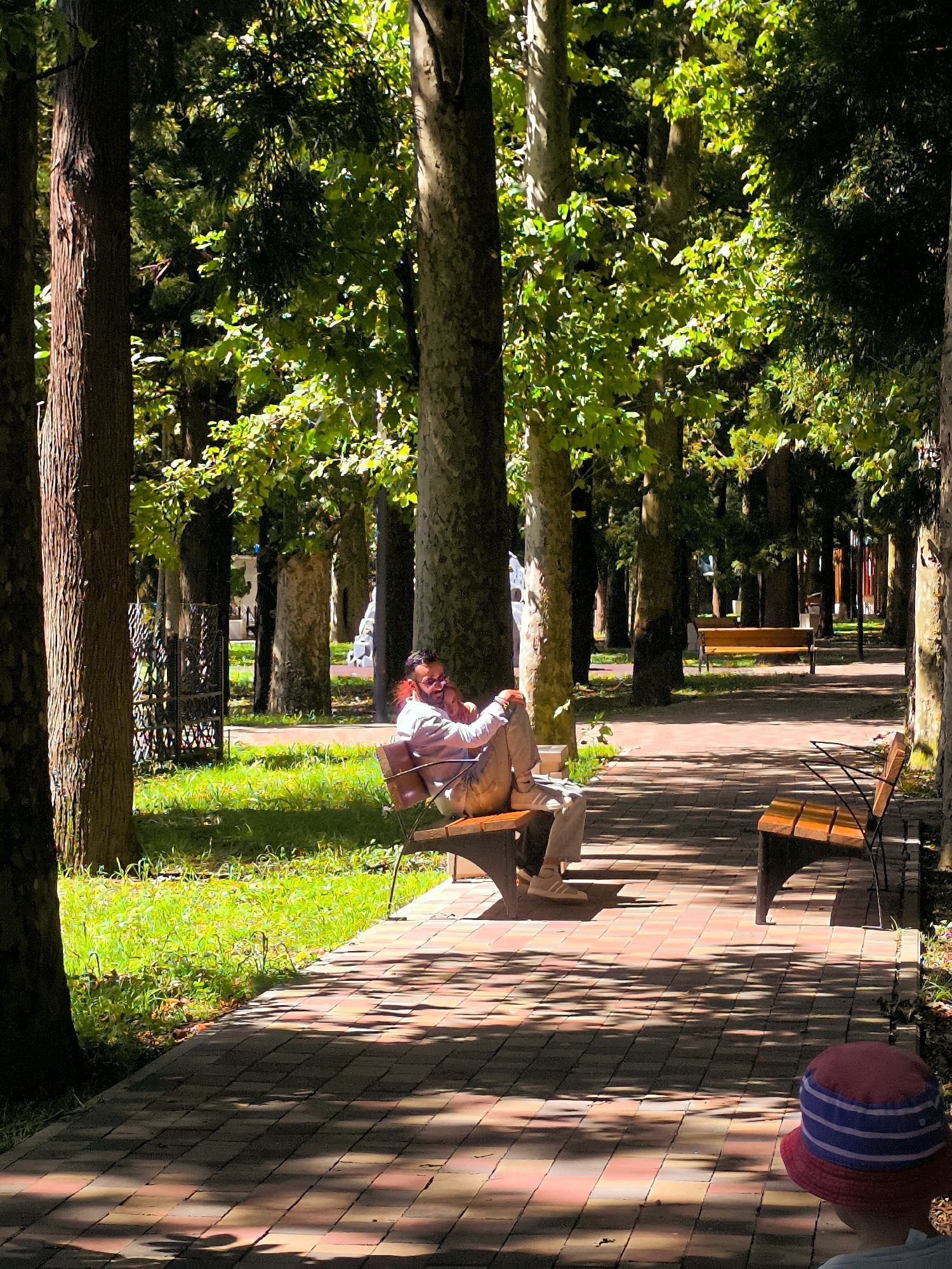 A person sits on a park bench in a tree-lined area. The person is wearing a light-colored shirt and light-colored pants, and is looking down. A red and blue striped hat is visible in the foreground. The ground is paved with bricks, and there is grass on either side of the path. Sunlight filters through the trees, casting shadows on the ground.