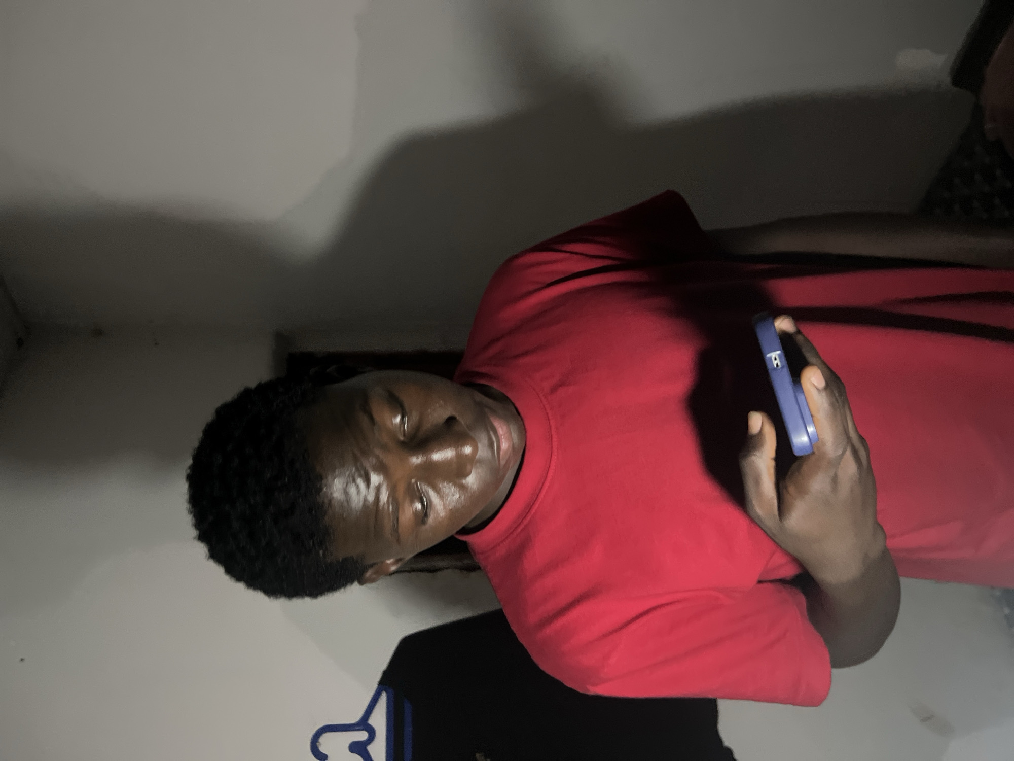 A young Black man is standing in a dimly lit room, wearing a red t-shirt. He is looking down at a blue smartphone he is holding in his right hand. A black t-shirt hangs on a blue hanger to his left. The wall behind him is plain and light-colored, with shadows cast upon it. The overall impression is of an indoor, domestic setting, likely a bedroom or living space, in Agwala, Nigeria.