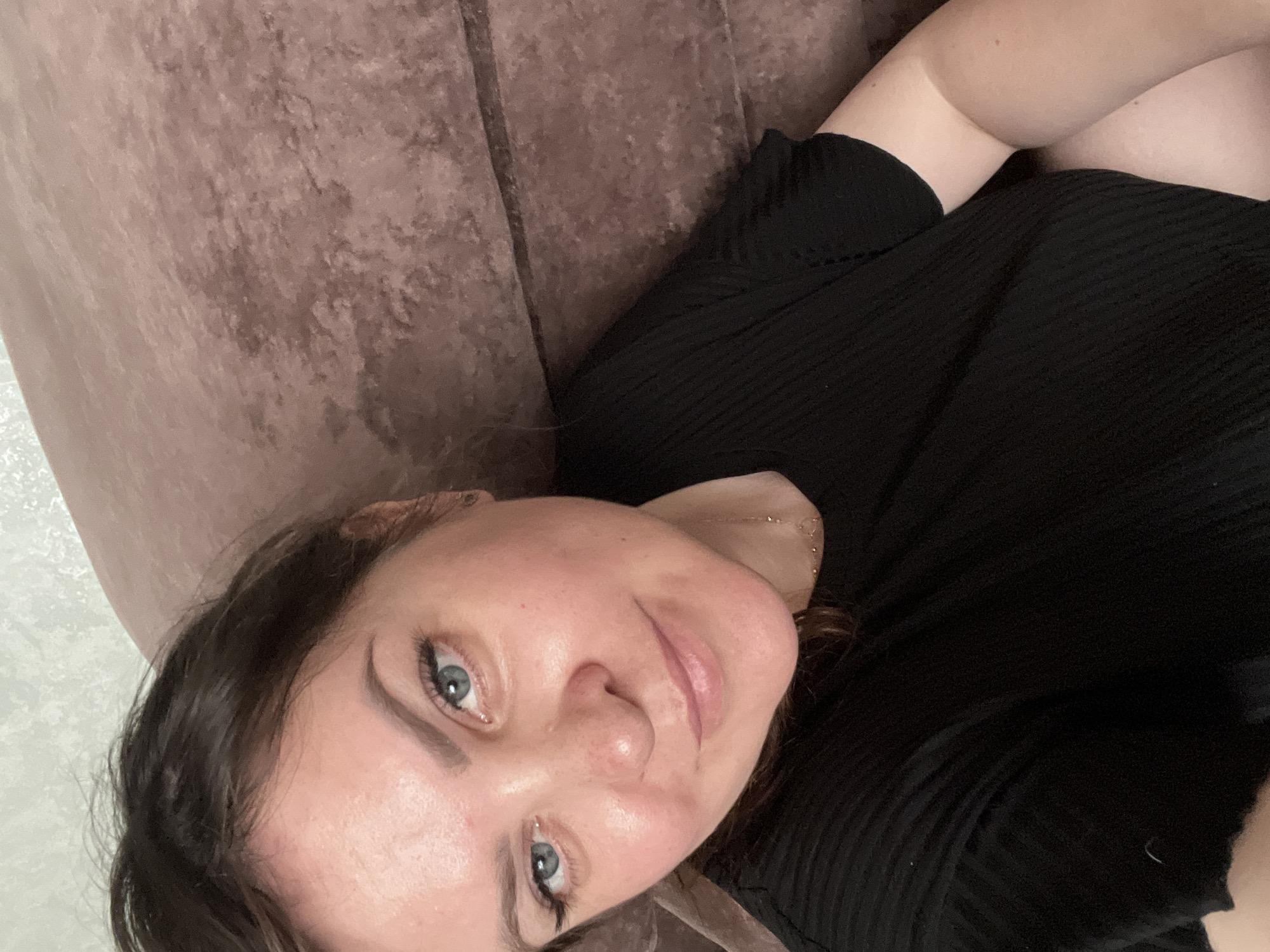 A woman is lying on a brown, textured couch or cushion. She is wearing a black, ribbed shirt. Her eyes are blue, and she has dark hair. She is likely indoors. There is no specific information in the image to confirm a location within Noviy, Russia, or any other city.
