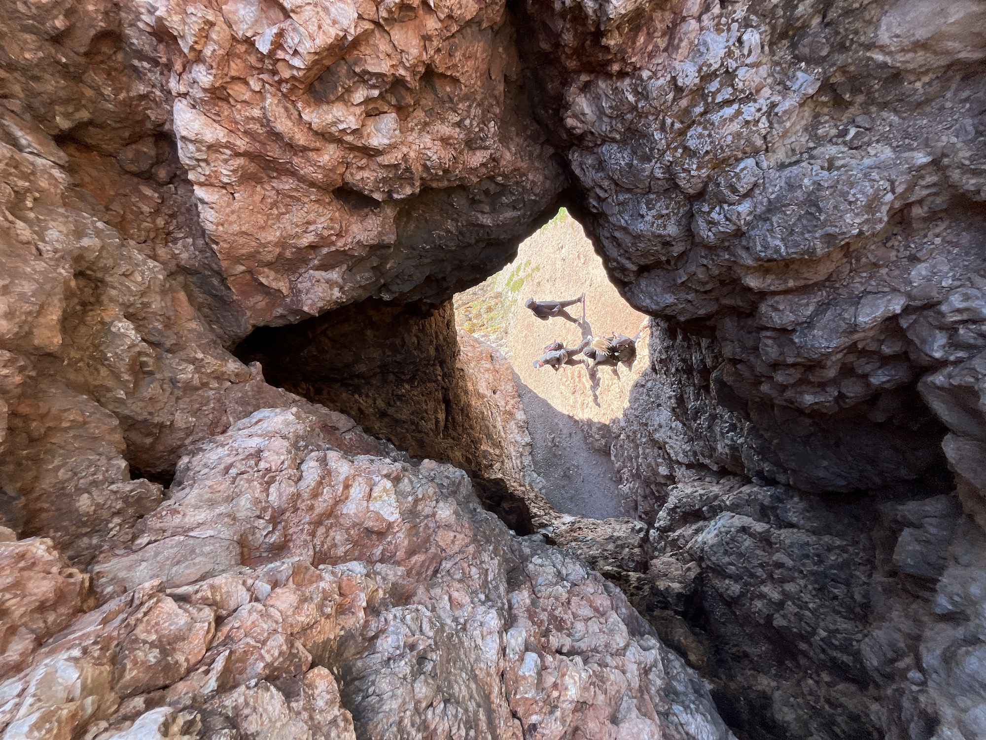 A view through a natural rock opening in Krasnoyarsk, Russia, reveals a bronze sculpture depicting a man atop several animals, located in an outdoor setting. The surrounding rock formations are rough and textured, with variations in color. The sculpture is positioned in direct sunlight, casting a shadow on the ground.