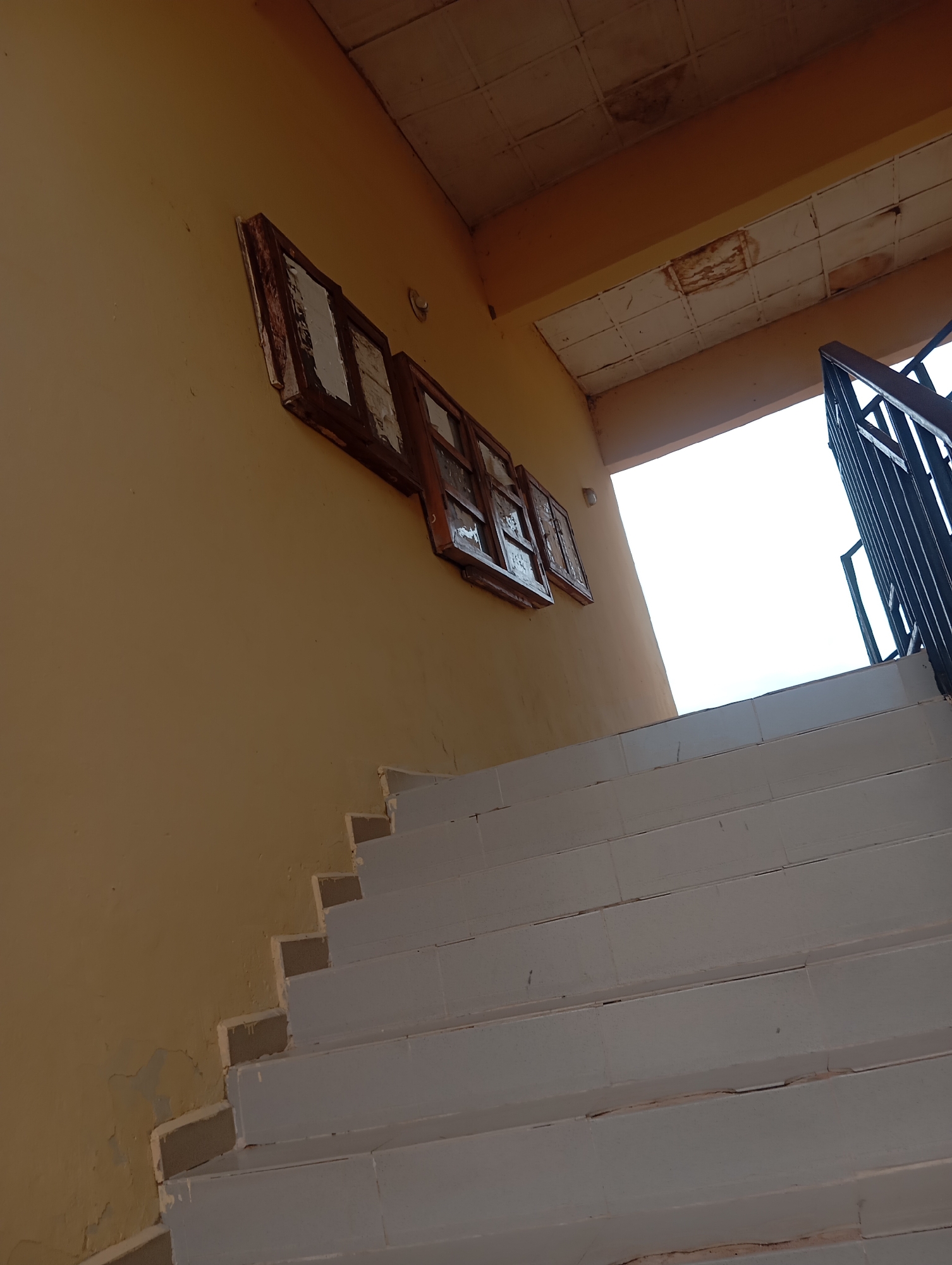 The image shows an outdoor staircase in Jalingo, Nigeria. The staircase is made of concrete and is painted white. The walls of the building are painted yellow. There are three windows on the wall above the staircase. The windows are made of wood and have a distressed appearance. The ceiling is made of tiles and appears to be damaged in places. A black metal railing runs along the side of the staircase. The sky is visible in the background and is a bright white, suggesting it is daytime. The overall scene depicts a somewhat neglected or weathered outdoor area.