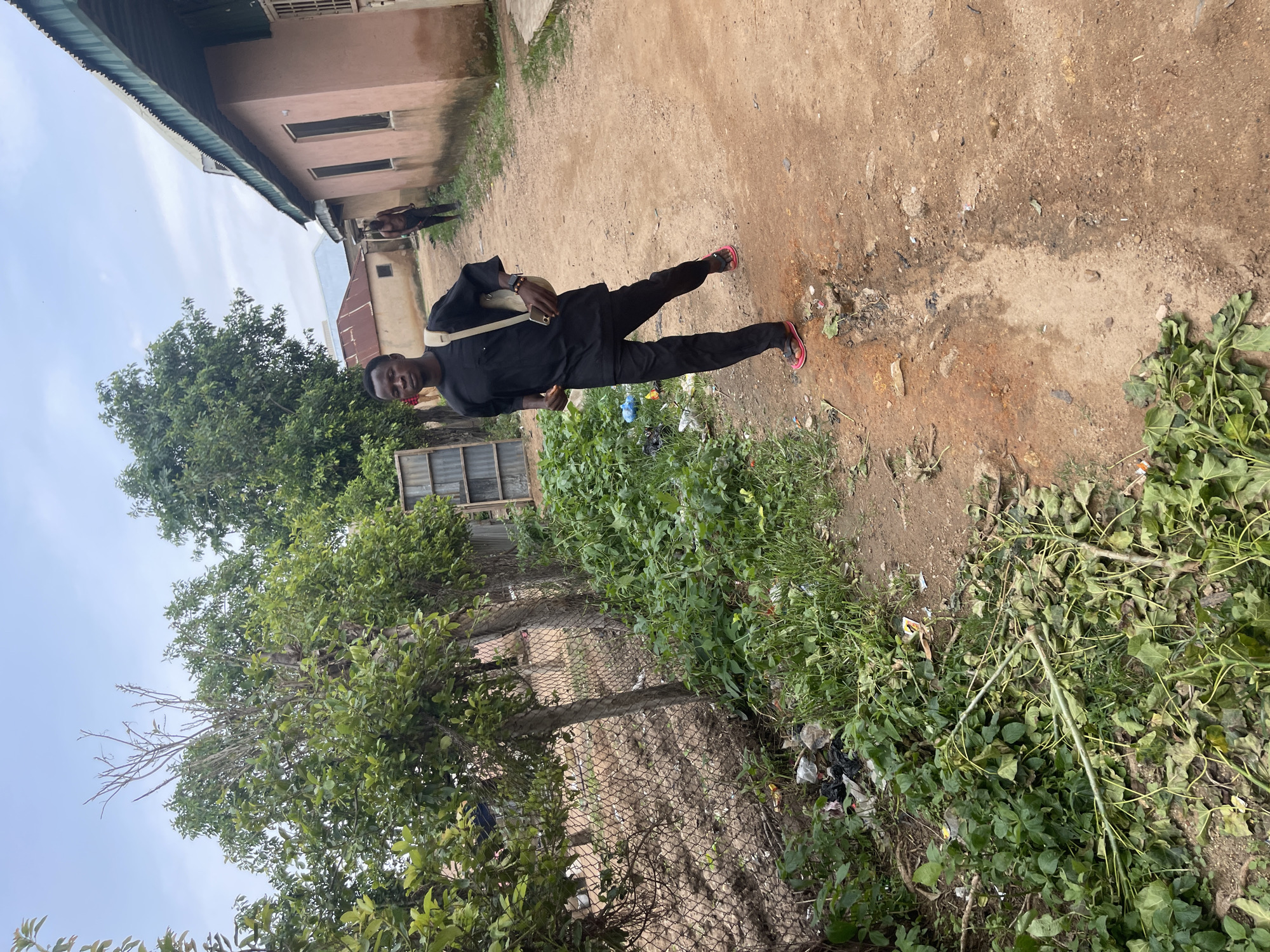 In Rafin Par, Nigeria, a young man wearing a black outfit and carrying a beige bag walks along a dusty, unpaved path. He is wearing red and black sandals. To his left, a chain-link fence borders an area with overgrown green vegetation, some of which has been cut and lies on the ground. Beyond the fence, trees are visible. To his right, a pale pink building with a corrugated metal roof runs alongside the path. Another man, shirtless and wearing dark shorts, is walking away from the camera in the distance, near the pink building. The sky is overcast with clouds. Debris, including plastic bags and leaves, is scattered along the side of the path.
