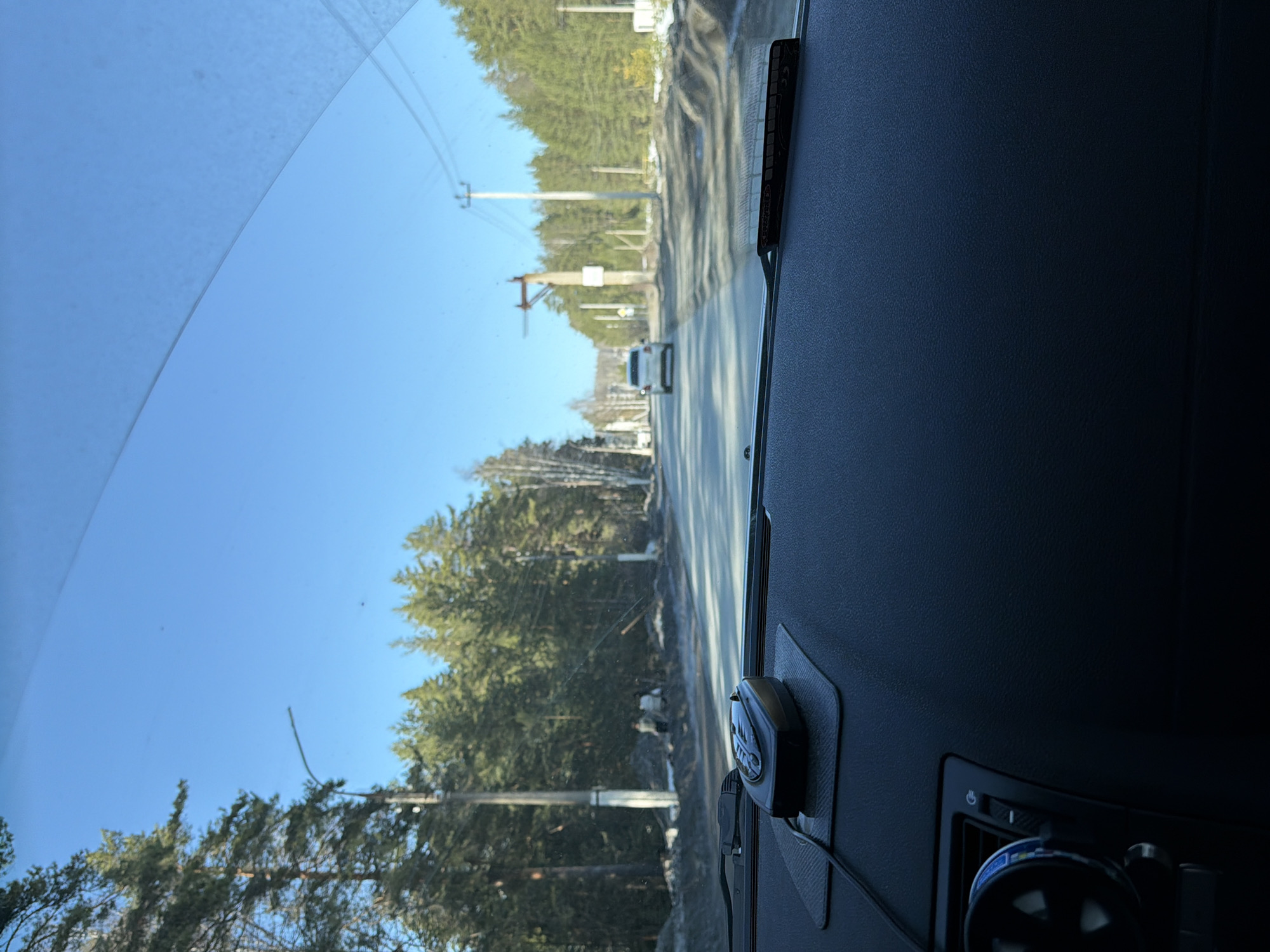 The image shows a view from inside a vehicle, likely a car, looking out through the windshield. The vehicle is on a road in Noviy, Russia. Outside the vehicle, the road is bordered by trees and power lines. There is a white vehicle in the distance on the road. The sky is clear and blue. The dashboard of the car is visible in the foreground, with a radar detector and an air vent visible.