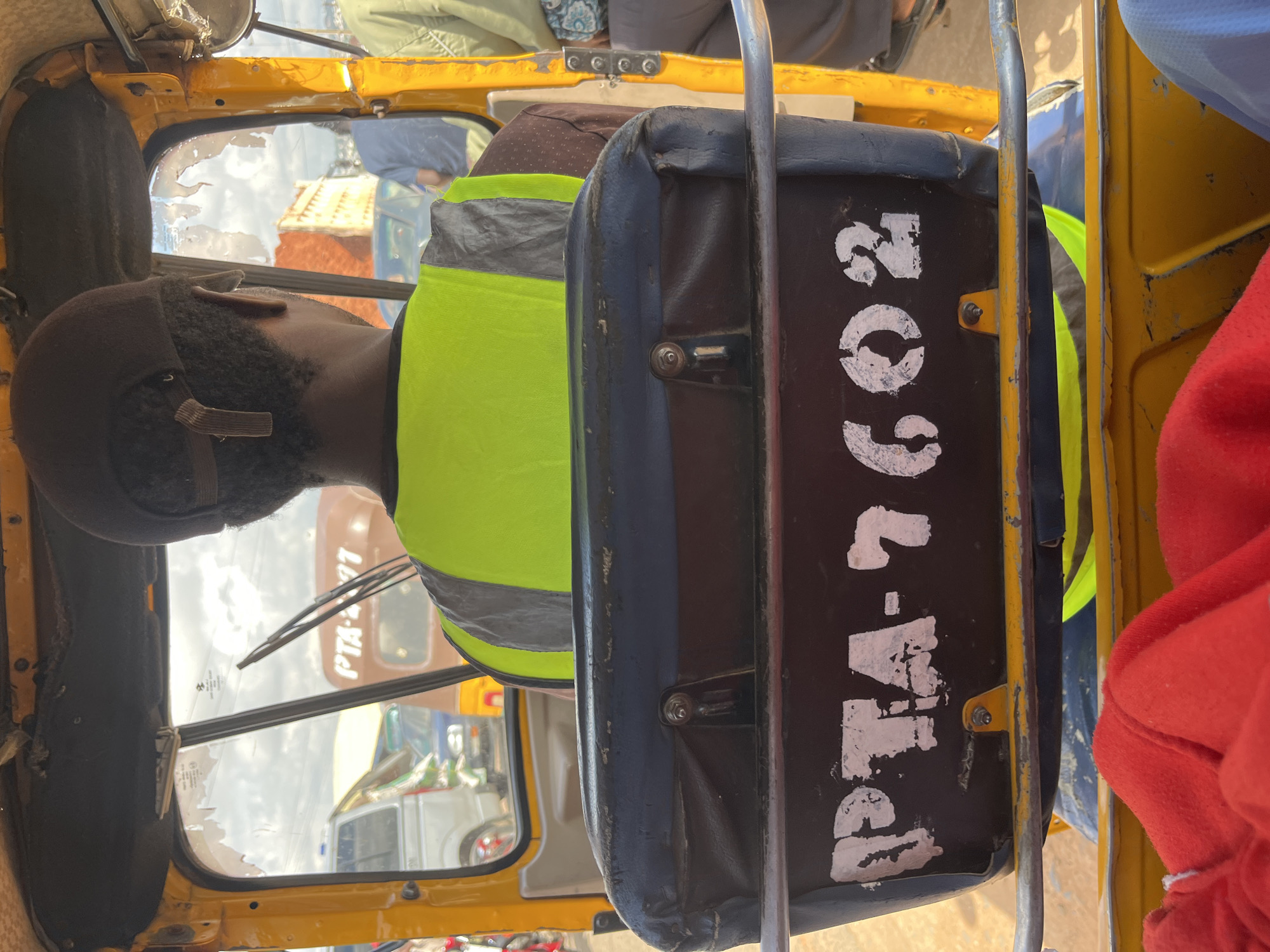 The image shows the interior of a yellow auto-rickshaw, commonly known as a "Keke Napep" in Nigeria. The driver, wearing a brown baseball cap and a bright yellow and grey reflective vest, is seen from behind, looking forward through the windshield. The windshield is cracked and dirty, with windshield wipers visible. Through the windshield, other vehicles and a building can be seen. A dark blue cushion with white stenciled text that reads "PTA-7602" is mounted behind the driver's seat. A red cloth is visible in the foreground, suggesting a passenger is seated in front of the camera. The overall impression is of a typical street scene in Jos, Nigeria, where such auto-rickshaws are a common mode of transportation.