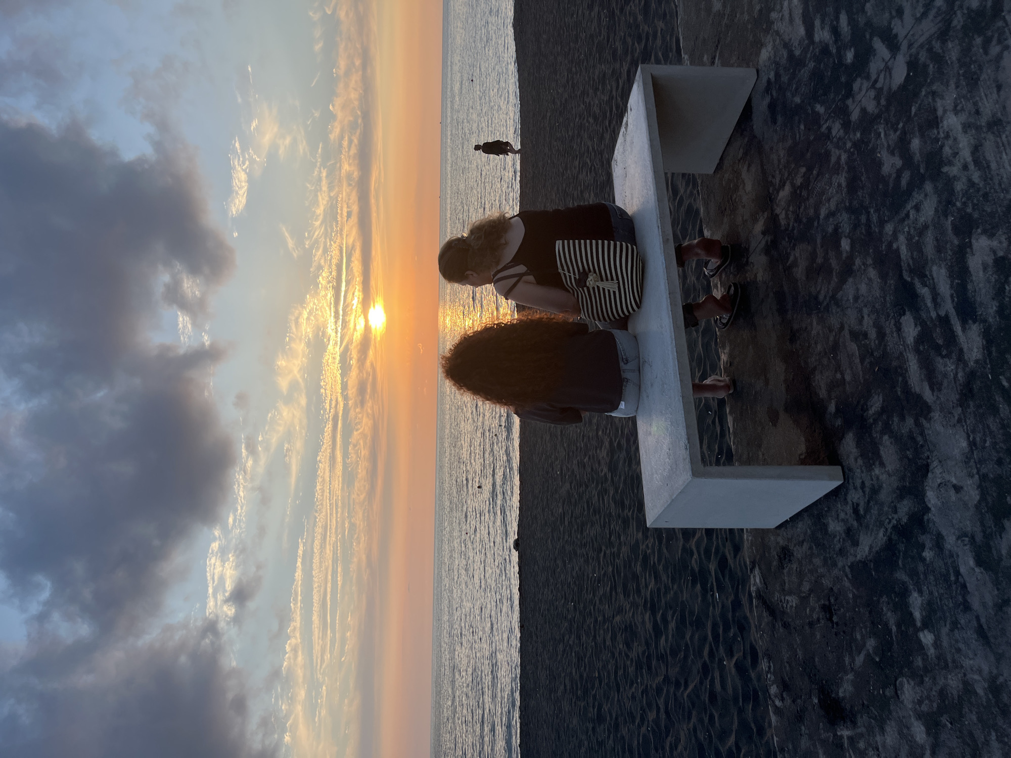 The image depicts a serene sunset scene on a beach in Ureki, Georgia. Two individuals are seated on a white concrete bench, facing the ocean. The person on the left has curly, dark hair and is wearing a dark top and jeans. The person on the right, wearing a black tank top and jeans, is holding a striped tote bag. Their feet, adorned with sandals, are visible beneath the bench. In the distance, a solitary figure walks along the shoreline, silhouetted against the vibrant orange and yellow hues of the setting sun. The sky is a canvas of colorful clouds, transitioning from deep blue to soft pastels. The beach itself is composed of dark sand, contrasting with the gentle waves of the Black Sea. The overall atmosphere is one of tranquility and natural beauty.
