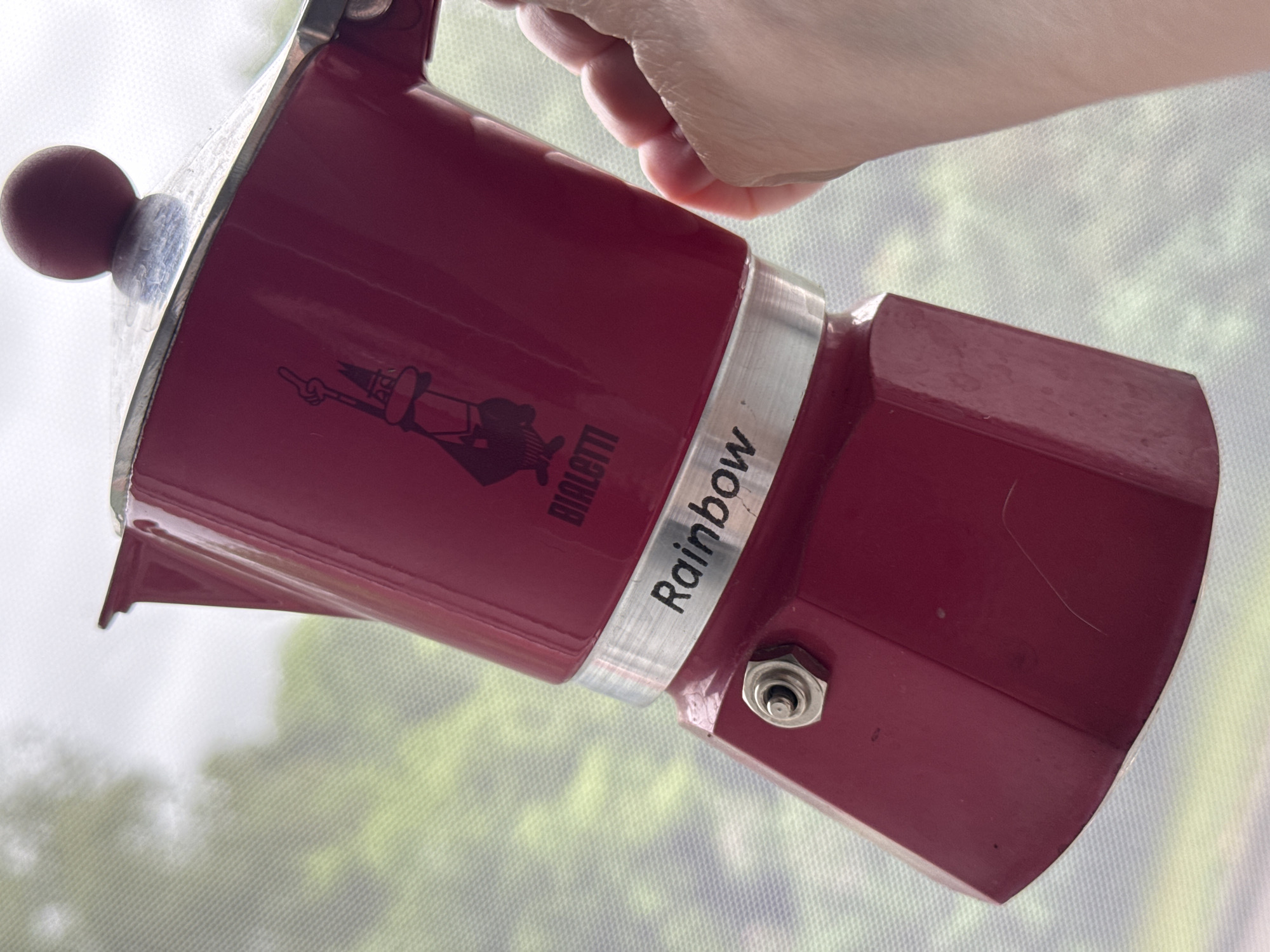 A red Bialetti "Rainbow" Moka pot is depicted at a slight angle, held by a human hand positioned on its right side. The pot features a hexagonal lower chamber with a silver pressure relief valve, and an upper chamber displaying the black Bialetti "little man" logo and the brand name "BIALETTI." A silver metallic band separates the two main sections, inscribed with the word "Rainbow" in black text. The pot is topped with a silver lid and a spherical red knob. The background is out of focus, showing blurred green and white hues, consistent with outdoor foliage and sky, seen through a fine mesh screen or net curtain. The specific location in the city of Riga, Latvia, cannot be determined from the visual information presented in the image.