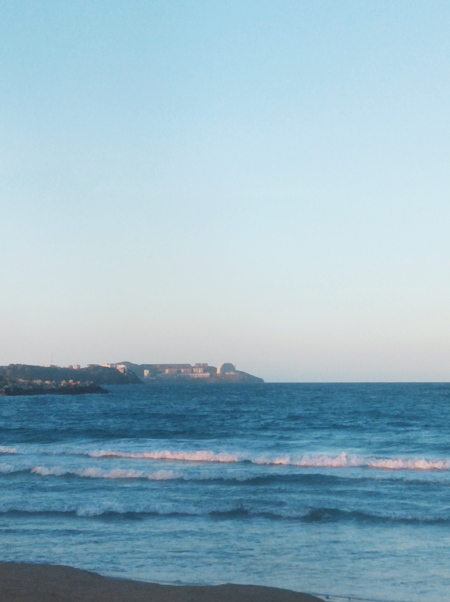 The image captures a serene coastal scene, likely at dusk or dawn, in Porlamar, Venezuela. The foreground shows a sandy beach. The ocean's surface has gentle waves with foamy crests. The sky above transitions from light blue near the horizon to a lighter hue overhead. In the middle ground, land with structures (buildings) is visible, likely a portion of the city.