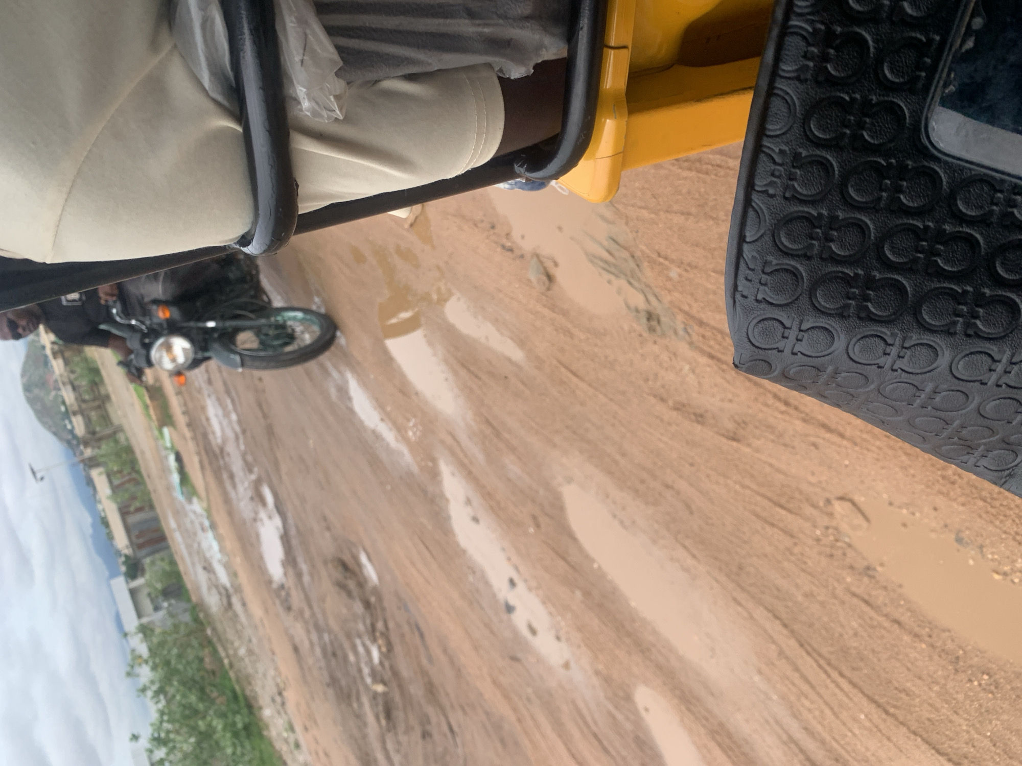 The image depicts a muddy dirt road in Jalingo, Nigeria, with puddles of water scattered across its surface. In the background, buildings and vegetation can be seen, with hills and a cloudy sky in the distance. A person is riding a motorcycle on the road, while the foreground shows a portion of a yellow vehicle, possibly a tricycle or a tuk-tuk, and a black bag with a patterned surface. The scene suggests a rural or semi-urban environment with unpaved roads, common in many parts of Nigeria, including Jalingo. The presence of the motorcycle and the tricycle suggests a mode of transportation. The weather appears overcast, contributing to the muddy conditions of the road.