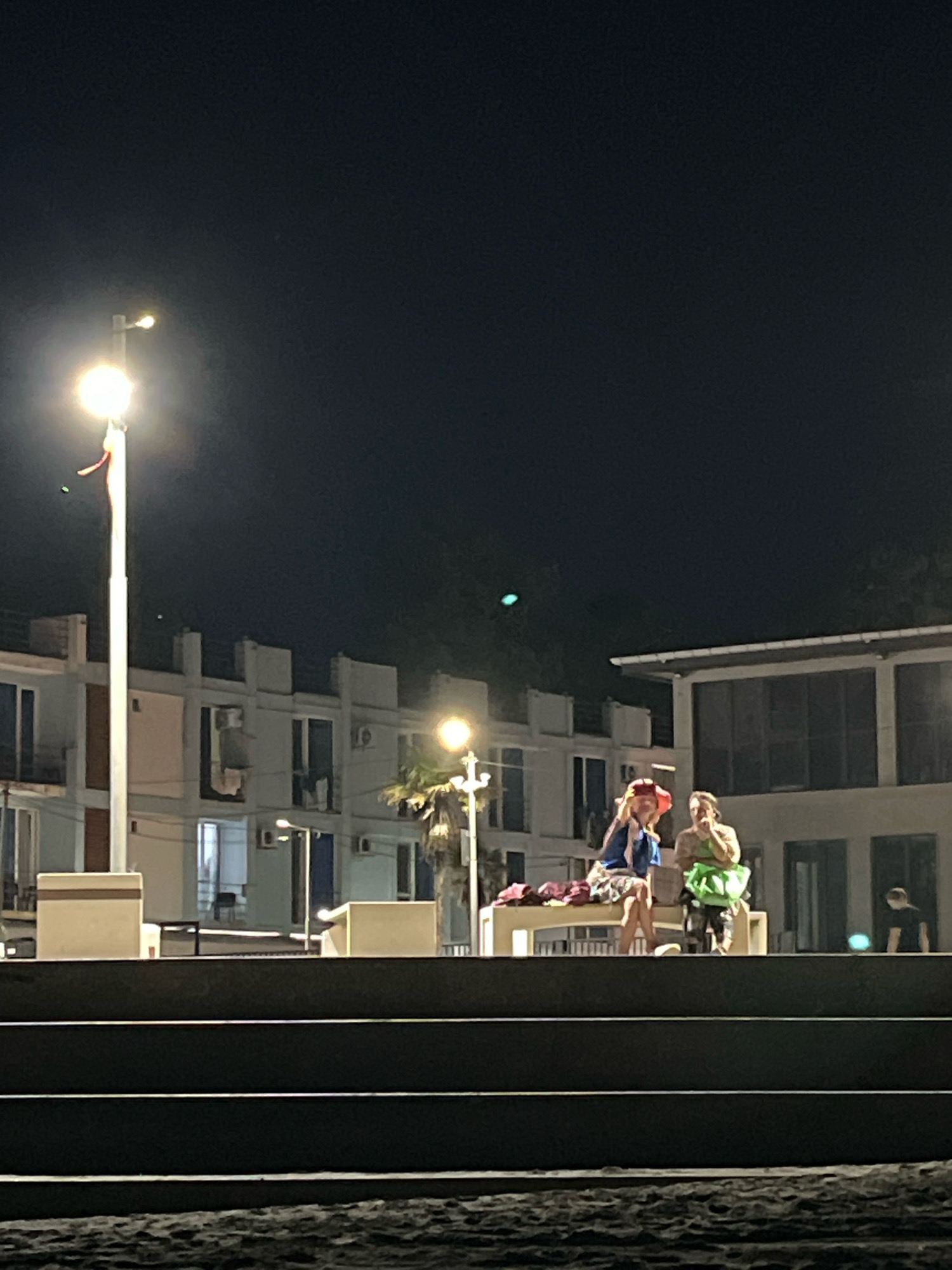 In Ureki, Georgia, at night, a wide shot captures two women and a man near a beach. In the foreground, dark, textured steps lead down to a sandy, wet area. The middle ground features a raised platform with a white bench. On the bench, a woman in a red hat and a blue jacket is seated, gesturing with her hands, while another woman in a green outfit stands beside her. To the right, a man in a black shirt stands in the background. Behind the platform, a multi-story building with white walls and dark windows is illuminated. A lamppost stands prominently to the left of the frame. Palm trees are visible to the right of the building.