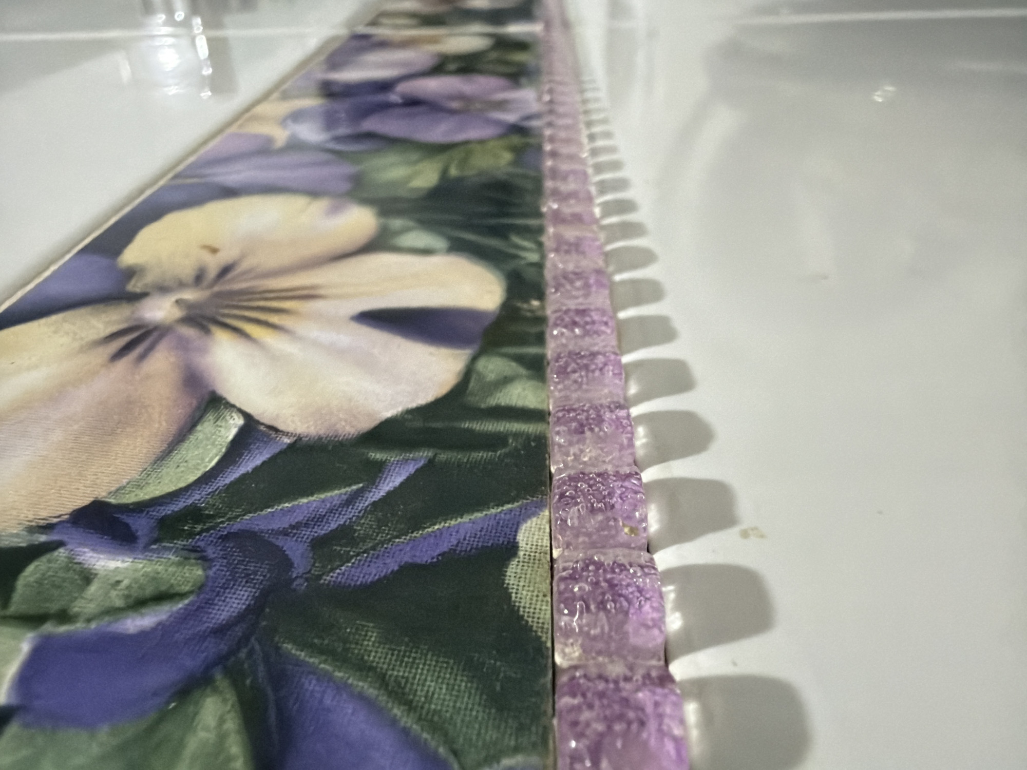 The image shows a close-up of a decorative border with a floral pattern. The border features an image of purple and yellow pansies with green foliage. Below the floral border is a row of small, translucent purple tiles with an iridescent quality, casting shadows on a white surface. The overall scene suggests a decorative element, possibly in a bathroom or kitchen backsplash. The image is taken from a low angle, looking up at the border. There are no people or animals visible in the image. There is no information that would indicate the location in Qarağandı city administration, Kazakhstan.