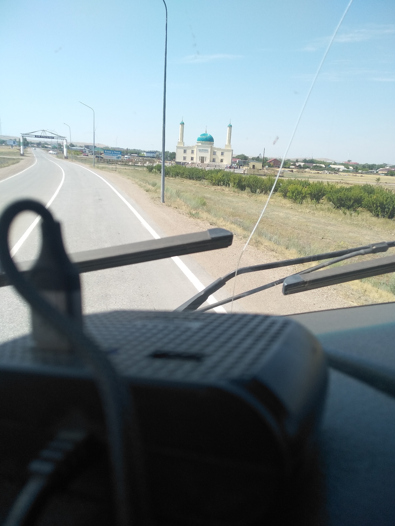 The image, captured from the interior of a vehicle, shows a road leading towards a town in Osakarovka, Kazakhstan. A mosque with a blue dome and a tall minaret is visible in the distance. The road is flanked by dry grass and scattered trees, with houses and other buildings forming the edge of the town. An archway with text is positioned over the road at the entrance to the town. The foreground of the image is dominated by the dashboard of the vehicle, including a black electronic device and a rearview mirror with wipers. The sky is clear and blue.
