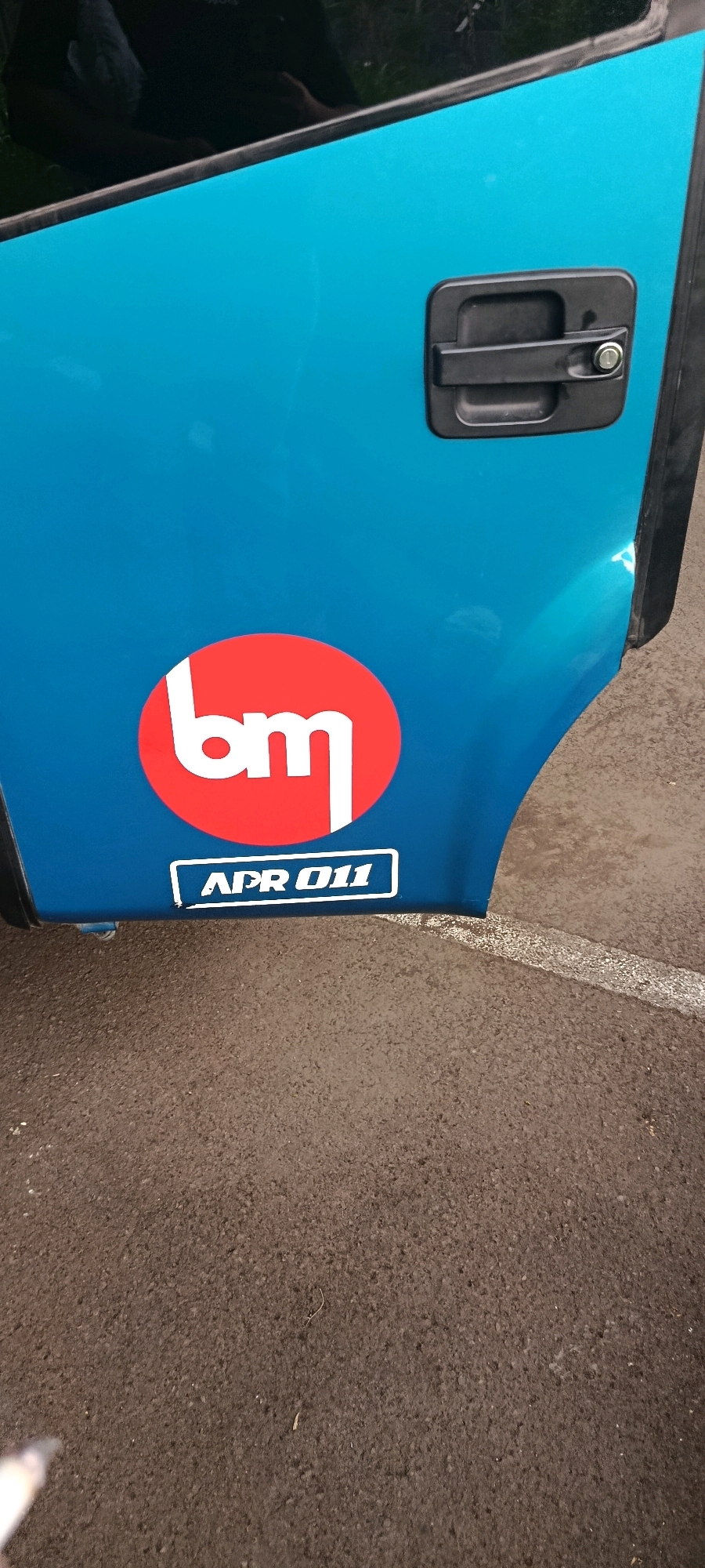 A close-up shot shows a blue door with two stickers. One sticker is a red circle with white letters that appear to spell "bm." Below that, a white rectangular sticker has the text "APR 011" in black letters. A black door handle is visible in the upper right corner of the frame. The door is part of a vehicle, likely a van or truck, and is parked on asphalt. A white line is visible on the asphalt in the lower right portion of the image. The background shows a reflection of a person in the upper left of the vehicle door. The scene appears to be outdoors, in a relatively open area.