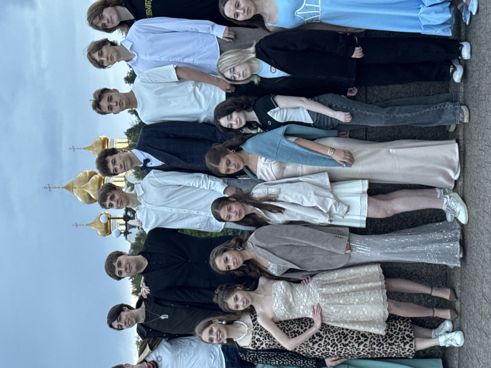 This media file shows a group of people, a mix of men and women, lying down in a line. The people are arranged vertically and appear to be looking up towards the sky. The setting includes what looks like the gilded domes of a church or cathedral in the background, suggesting a location of cultural or religious significance. The weather appears overcast, with a soft, diffused light.
The people are dressed in a variety of clothing. Some men are wearing shirts and jackets, while others are in more casual attire. The women are wearing dresses, ranging from formal to more casual styles, some with sequins and lace. Their expressions are varied, with some appearing calm or contemplative, while others have smiles on their faces. The overall impression is that of a staged photograph or a moment of posed relaxation among a group of friends or participants in an event.
There is no visible text in the image. The time of day seems to be daytime, given the overall lighting. The arrangement of the people and the architectural elements in the background suggest a possibly European setting, though this cannot be definitively determined. The activity appears to be a posed group shot, with the individuals lying down side-by-side.