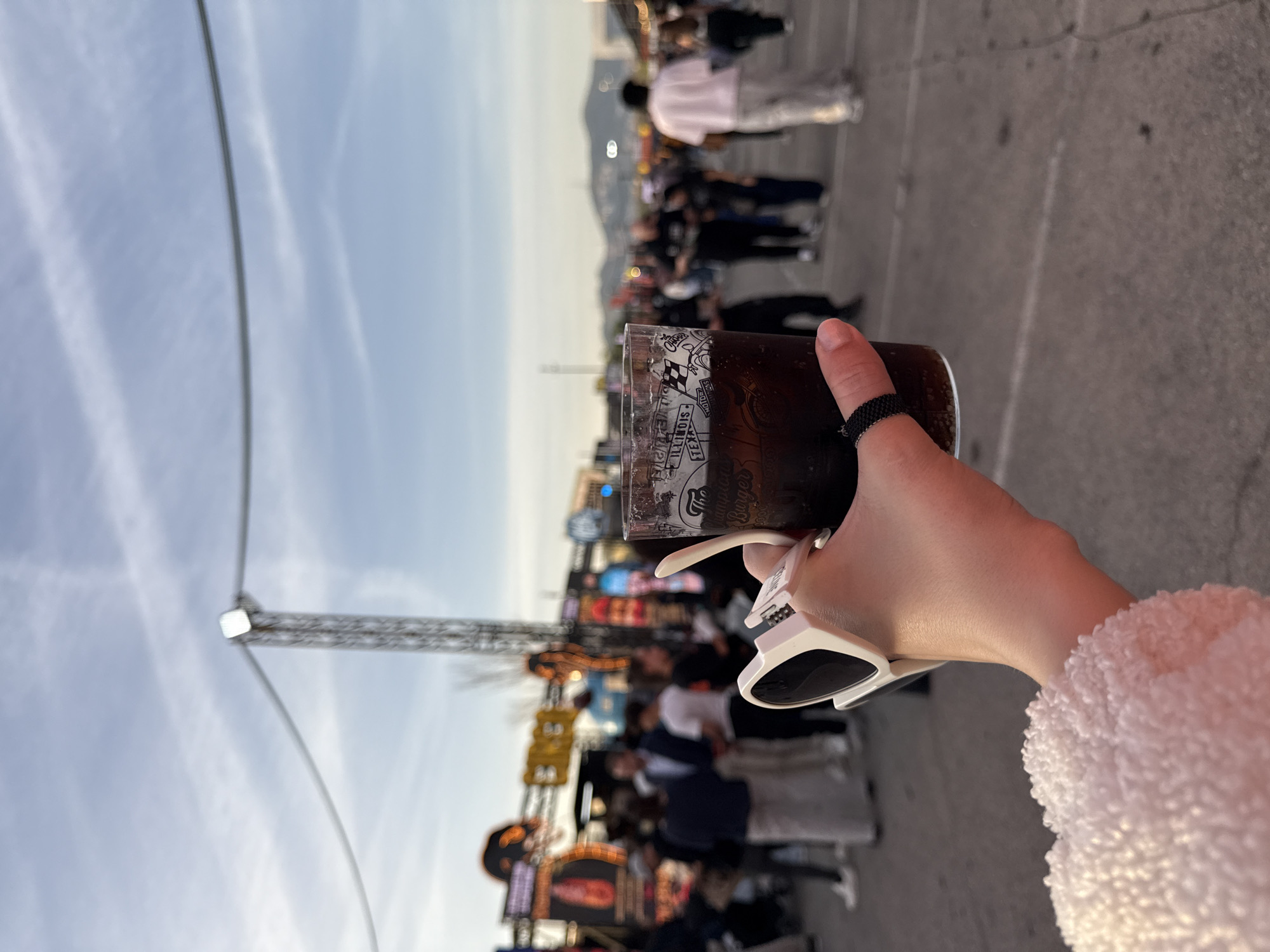 Here is a detailed description of the image:
**Scene:** The photograph captures a moment at what appears to be an outdoor event or fair in Barcelona, Spain. The image is oriented vertically.
**Foreground:** A person's hand is holding a glass of dark liquid, likely a soda or cola. The person is wearing a black beaded ring on their index finger. White sunglasses are clipped onto the side of the glass. The arm is covered in a fuzzy white fabric.
**Background:** Behind the hand and glass, there is a lively, bustling environment. There are numerous people walking around, many of whom appear to be families or groups. There are colorful signs and lights, suggesting it is an entertainment or festival area. Some signs are partly legible and appear to have "Burger" written on them, possibly indicating food stalls. The Ferris wheel scaffolding can also be seen.
**Environment:** The sky above is pale and slightly overcast. The ground appears to be a paved area, possibly concrete. The city skyline is visible in the distance.