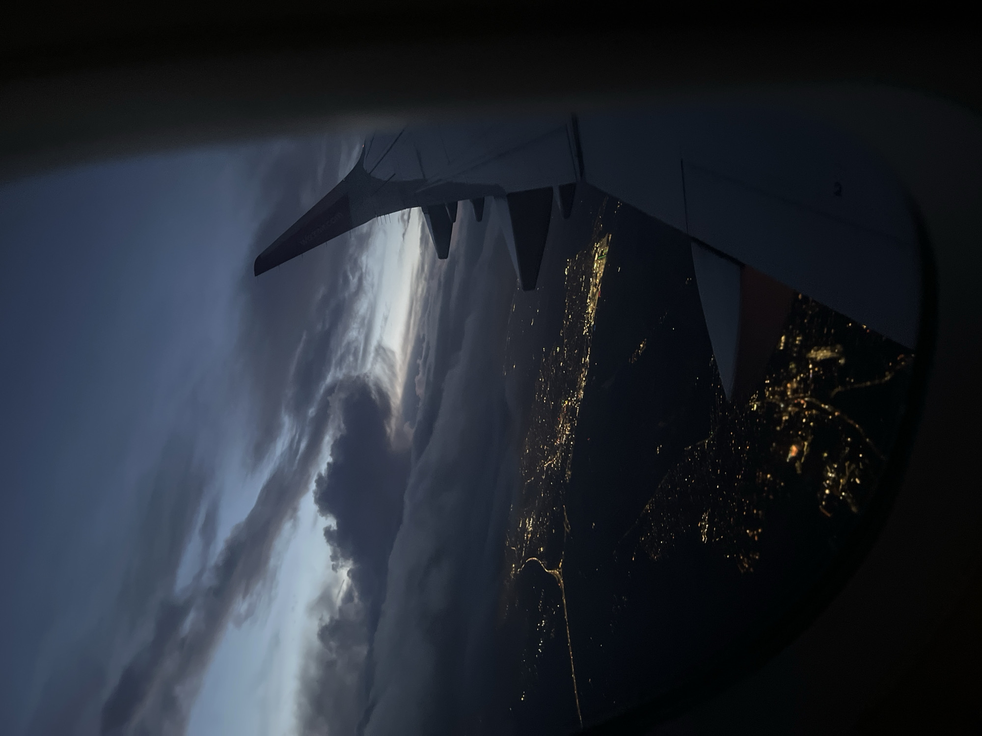 The image is a view from an airplane window. An airplane wing is visible in the upper portion of the frame. Below the wing, there are dense clouds on the left side and city lights on the right, presumably viewed at night. It is impossible to specify if the city is St Albans, United Kingdom, from this image. The city lights are concentrated and appear to indicate urban development.