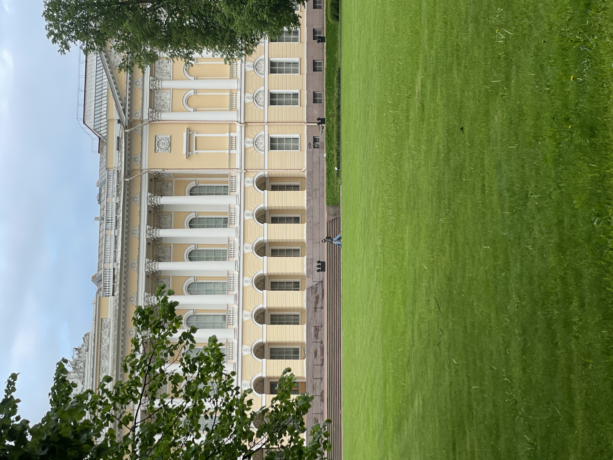 The image shows a building with a cream-colored facade and a green lawn in front of it. The building has multiple windows, white columns, and intricate decorations. A tree with green leaves is partially visible in the foreground. A person can be seen walking along the edge of the lawn. The location is in an urban area, as indicated by the architectural style of the building.