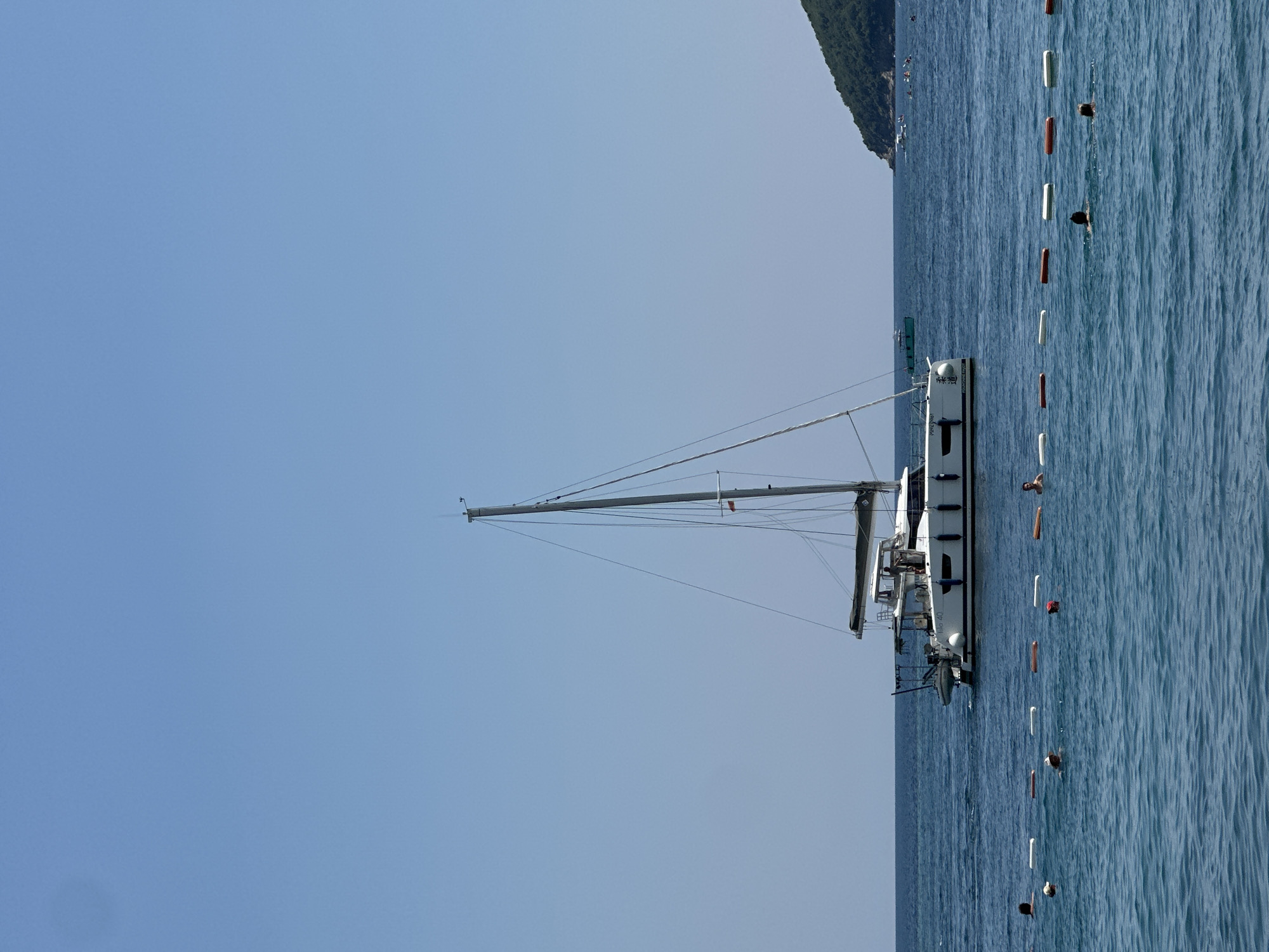 This image shows a sailboat docked in the water near the coast of Budva, Montenegro. The sky is a clear, pale blue, suggesting a sunny day. The water is a deep blue, with gentle ripples reflecting the sunlight. A line of buoys, alternating between white and orange, marks a boundary in the water. Several people are visible in the water, swimming or wading near the buoys. The sailboat is white with a tall mast and rigging. The hull of the boat is visible, as is some of its superstructure. In the background, a green, tree-covered hill rises from the coastline, characteristic of the Montenegrin coast. The image appears to be taken during daylight hours. The overall scene conveys a sense of a peaceful day at the beach or sea.