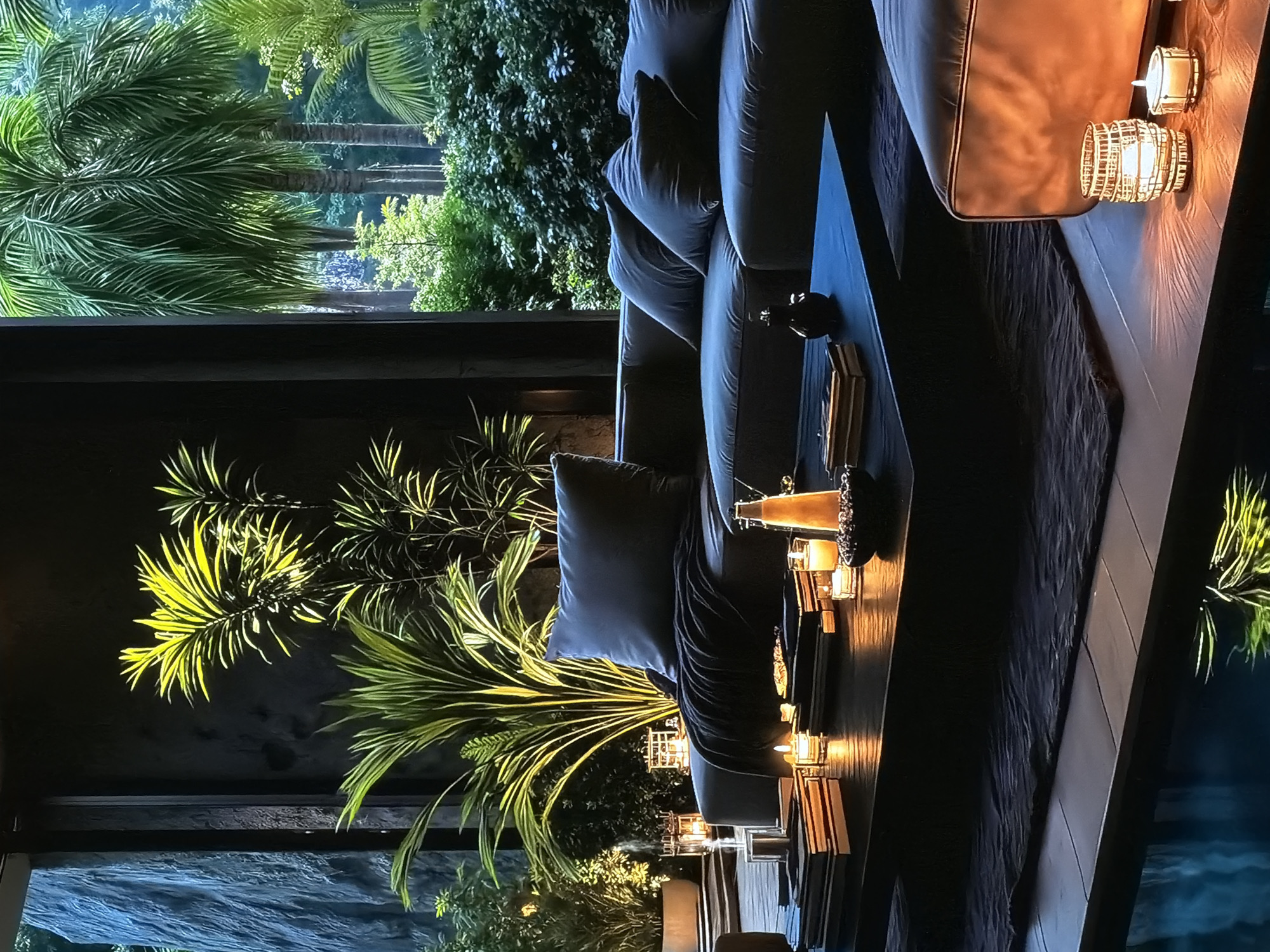 The image depicts a luxurious and tranquil indoor-outdoor lounge area, likely in the evening or at night. The scene is presented vertically, but mentally rotating it counter-clockwise reveals a horizontal view of a sophisticated living space overlooking lush tropical foliage.
In the foreground (left side of the mentally re-oriented image), a comfortable, dark-colored sofa with throw pillows is visible. Adjacent to it is a long, dark coffee or console table adorned with numerous decorative candles in various holders, including some with a wicker-like texture and others made of glass. The warm, flickering light from these candles casts a cozy glow on the table and surrounding area, suggesting an inviting and relaxed ambiance. A dark throw blanket is draped over the sofa.
Through a large window or open partition (right side of the mentally re-oriented image), a vibrant outdoor garden is visible. Several tall palm trees with prominent, feathery fronds are brightly illuminated by artificial lighting, showcasing their vivid green and yellow hues against the dark background of the night. Other dense green foliage is also present, with hints of blue in the lower right corner suggesting a body of water, possibly a swimming pool or pond, reflecting the night sky or artificial lights.
There are no people visible, and no specific activity is taking place, but the setup strongly suggests a space for relaxation and enjoyment. The contrast between the dim, warmly lit interior and the dramatically illuminated tropical exterior creates a serene and exotic atmosphere. No visible text is present in the image. The location is Sofiivska Borshchahivka, Ukraine.