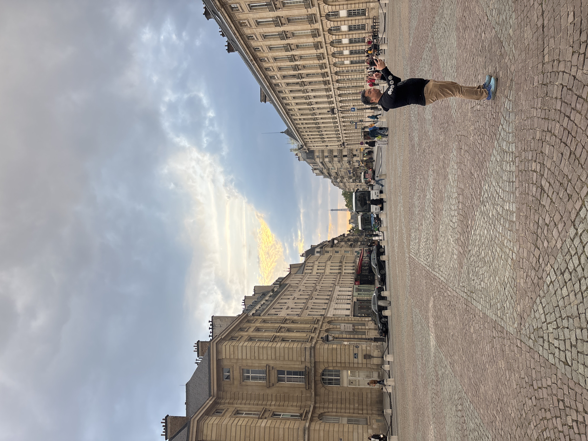 A medium eye-level shot depicts a street scene in Paris, France. The Eiffel Tower is visible in the distance. A man stands on the right side of the frame, holding a camera. He wears a black jacket, khaki pants, and blue sneakers. The street is paved with cobblestones. Tall buildings line both sides of the street. Cars are parked along the sides. The sky is cloudy, with some golden light near the horizon. Other people can be seen further down the street.