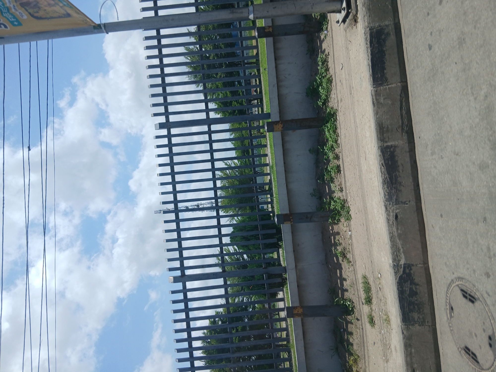The image shows a metal fence running along a sidewalk. Behind the fence, there's a green area with trees and possibly vehicles. The sidewalk has a concrete curb and a manhole cover. The sky is visible through the fence, featuring blue sky and white clouds. Power lines are present in the upper left portion of the frame.
Given the specification of Animashawun, Nigeria, and the general urban appearance, the location is plausibly a public area, such as a park or a commercial area, within Animashawun, Nigeria, although specific landmarks are not visible to confirm the exact spot.