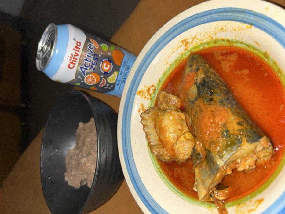 An indoor scene in Egbe, Nigeria, displays a prepared meal on a brown wooden surface. In the foreground, a white ceramic plate with blue and green concentric rings on its rim contains a bright red, oil-rich stew with one large piece of fish and one smaller, irregularly shaped piece of protein. To the left, a black ceramic bowl holds a quantity of light brown, lumpy food paste. Behind the plate, an aluminum can of Chivita Active Zest mixed fruit sparkling drink, featuring a light blue and white label with citrus fruit graphics and "Enriched with Vitamin C" text, stands upright with its pull-tab opened. The background is dark and out of focus. No individuals are present.