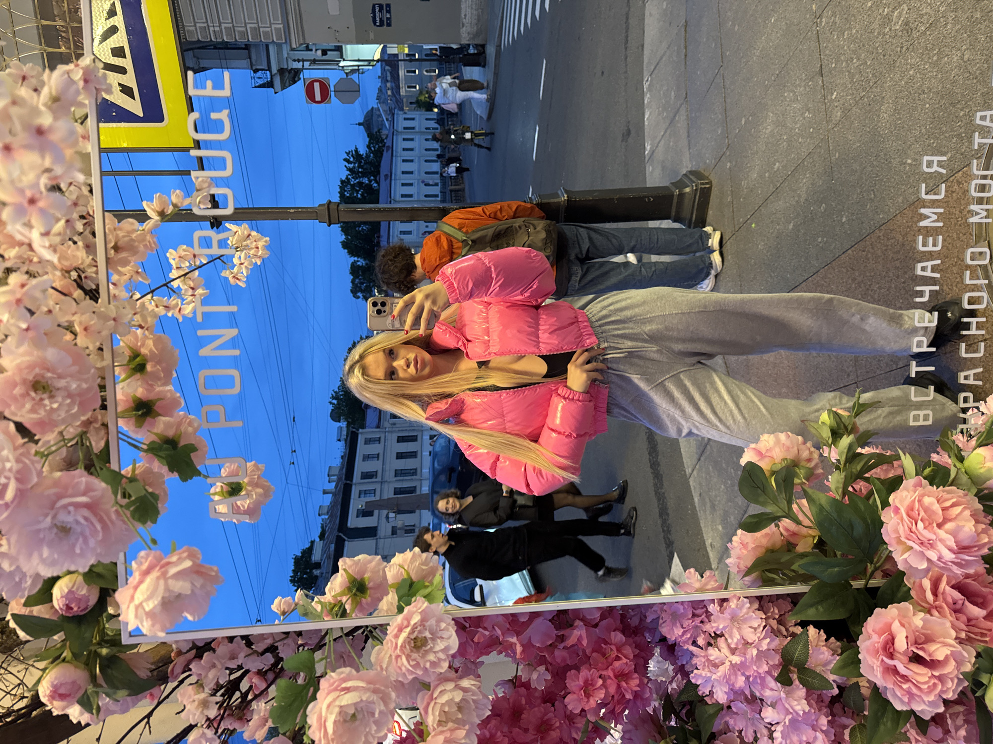 In this vertical, reflective image, a young woman with blonde hair poses for a selfie, holding her phone in her right hand. She is wearing a bright pink, puffy jacket and grey sweatpants. The setting appears to be a street in Saint Petersburg, Russia, as suggested by the visible text and architectural style. The time of day is dusk, with a twilight sky visible in the reflection.
The reflection shows a street scene with people walking, a crosswalk, and buildings in the background. A prominent sign reflected in a mirrored surface reads "PON ROUGE" in large, white letters. Another line of text in the reflection, also in white letters, appears to be in Russian and says "ВСТРЕЧАЕМ У СМОЛЬНОГО МОСТА" which translates to "MEETING AT SMOLNY BRIDGE." The foreground is adorned with artificial pink and white flowers, adding a decorative element to the scene. The overall mood is casual and youthful, captured during a moment of outdoor leisure.