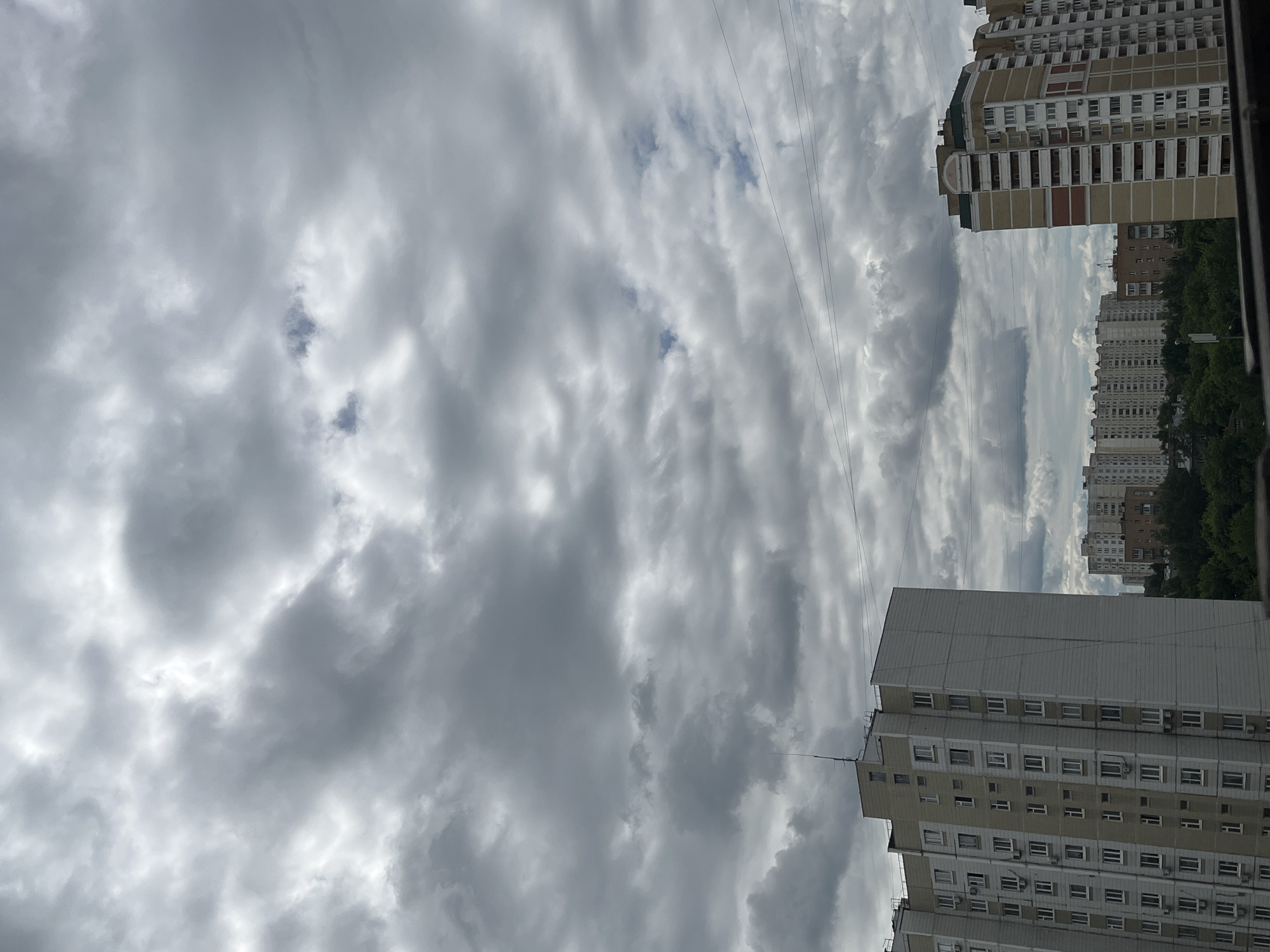 The image shows a cloudy sky over a cityscape in Moscow, Russia. The foreground features two tall residential buildings, one on the left and another on the right. The building on the left is lighter in color, with a grey and beige facade, and many windows. The building on the right is taller and has a more complex design with yellow, white, and red sections. In the background, more residential buildings and lush green trees are visible. Power lines crisscross the sky above the buildings. The clouds are dense and grey, suggesting an overcast day, with some patches of lighter sky peeking through. There are no people or vehicles visible in the image. The scene appears to be a typical view of a residential district in a large city.