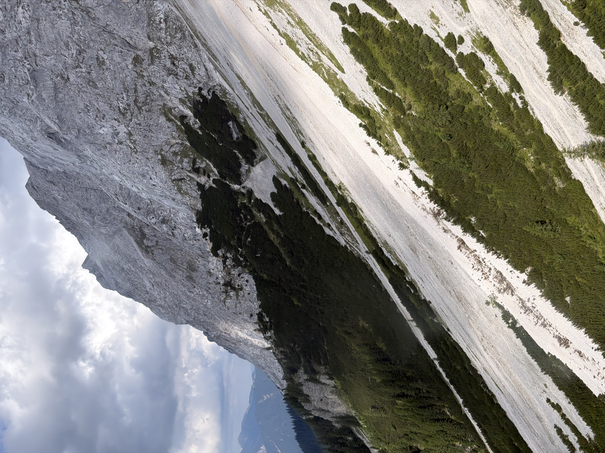 This is a photograph of a steep, rocky mountain slope in Grainau, Germany. The mountain is covered in scree and interspersed with patches of dark green coniferous trees. The sky is overcast with dramatic clouds. In the distance, other mountains are visible, partially obscured by haze. The image captures the rugged terrain and the transition from forest to barren rock.