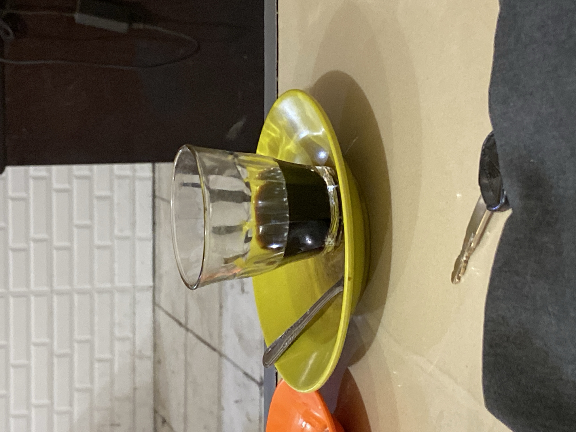 An overhead shot shows a shot glass filled with dark liquid on a bright yellow saucer. A silver spoon rests on the saucer, pointing towards the left. To the left of the saucer is an orange dish with a portion of its rim visible. In the foreground, a dark gray fabric is draped, and a car key with a black fob is placed on it. The background consists of a white brick wall with gray grout lines and a dark brown cabinet. The scene appears to be set on a light beige table. This image was likely taken in Lhokseumawe, Indonesia.