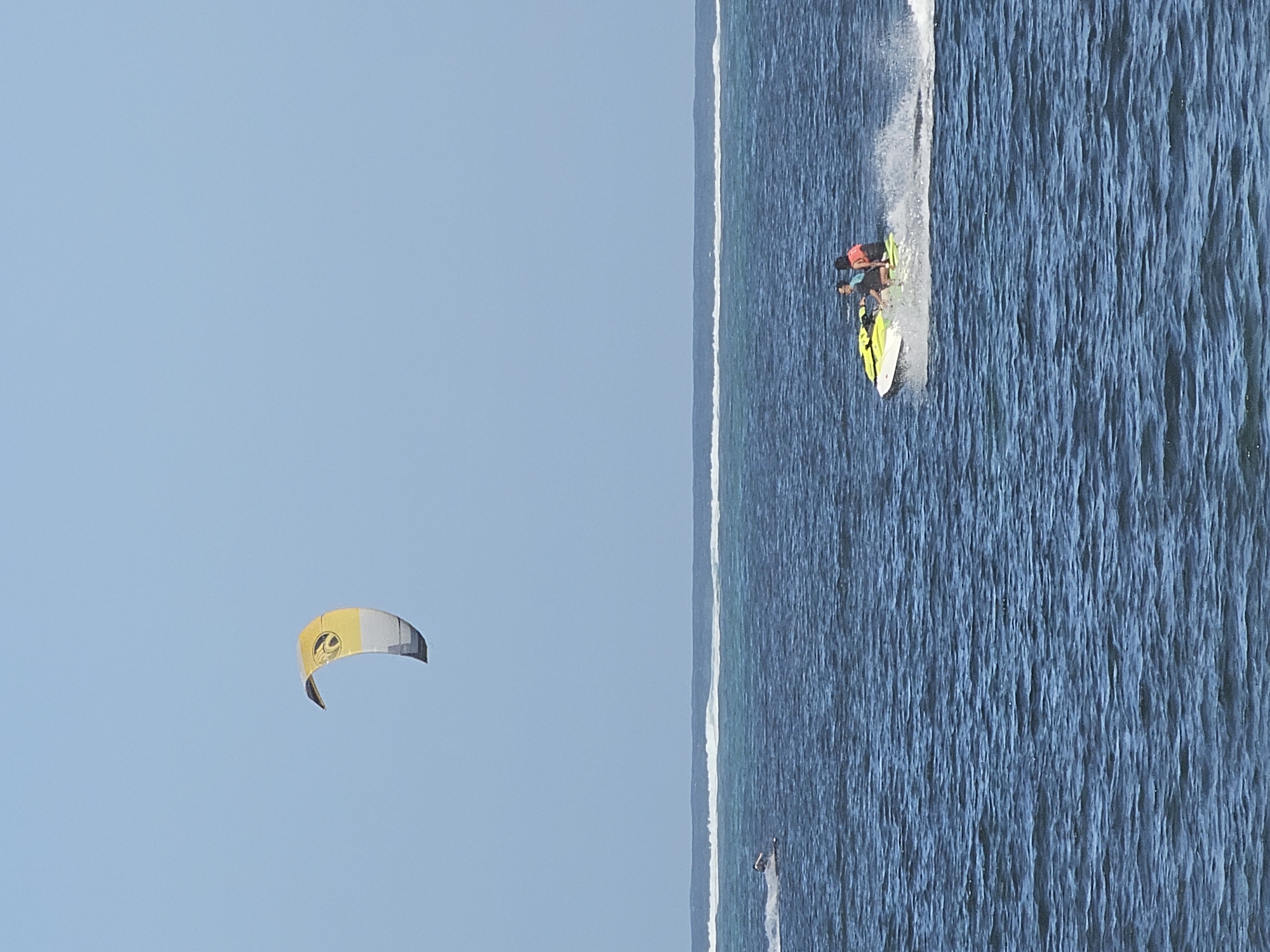 A slightly high-angle, medium-distance shot depicts a sunny beach scene. On the left of the frame, a yellow and grey kite soars in a clear blue sky. To the right, the blue ocean stretches, marked by visible wave patterns. A yellow jet ski with two people, one wearing an orange life vest, creates a white spray trail as it speeds along the water. In the distance, another individual appears to be surfing or kitesurfing. The image appears to be a coastal setting. The location is likely Sanur Beach, in Denpasar, Indonesia, due to its popular kitesurfing activity.