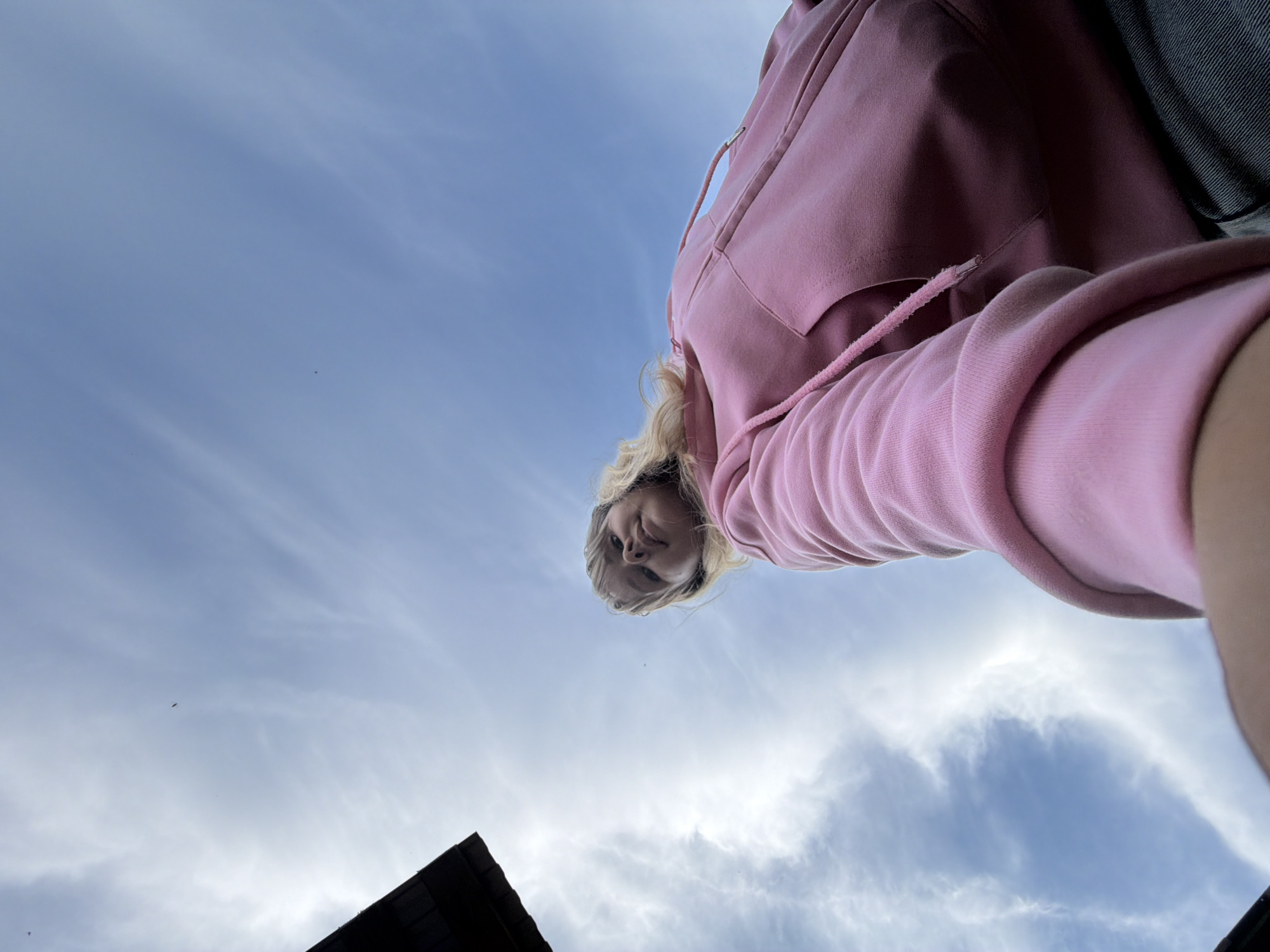 An upward-angle wide-shot photograph captures a young person from the chest up, looking down towards the camera, set against a predominantly blue and cloudy sky. The subject, who appears to have blonde hair, is wearing a pink hooded sweatshirt (hoodie) with visible drawstrings. Their face is seen from a low perspective, implying the camera was positioned below them. Part of the subject's arm is visible on the right side of the frame, and a dark strap or bag is partially seen on the far right edge.
The sky occupies most of the background, featuring light blue hues with scattered, wispy white clouds that suggest an overcast or partly cloudy daytime. In the bottom left corner, a dark, angled structure, possibly a roof or part of a building, is visible, adding a small architectural element to the otherwise expansive sky. No specific activity beyond posing for the photograph is evident. The image appears to be taken outdoors during daylight hours in Chumnaya, Russia. There is no visible text in the image.