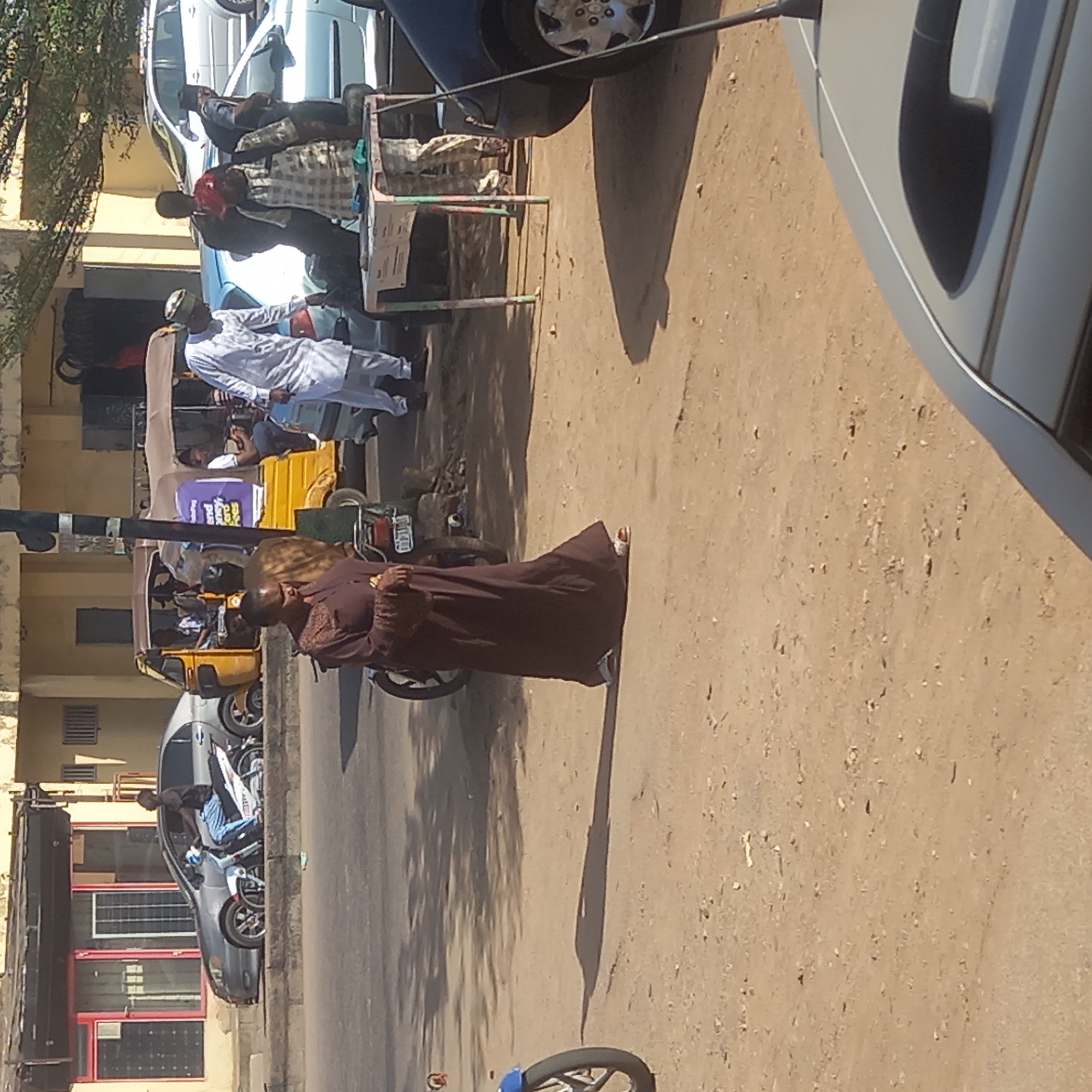 The image, taken in Sardauna, Nigeria, shows a street scene with several vehicles, pedestrians, and commercial activity. In the foreground, a woman wearing a long brown dress walks along the road. In the background, several yellow auto-rickshaws (tuk-tuks) are parked along the street. Several people are gathered around a table covered with goods, likely a vendor selling items. A man in a white robe stands nearby. Cars are parked along the side of the road, with one vehicle partly visible in the foreground. The street surface is dirt and uneven. Buildings are visible on the opposite side of the street, and tree branches appear in the upper left corner of the image.