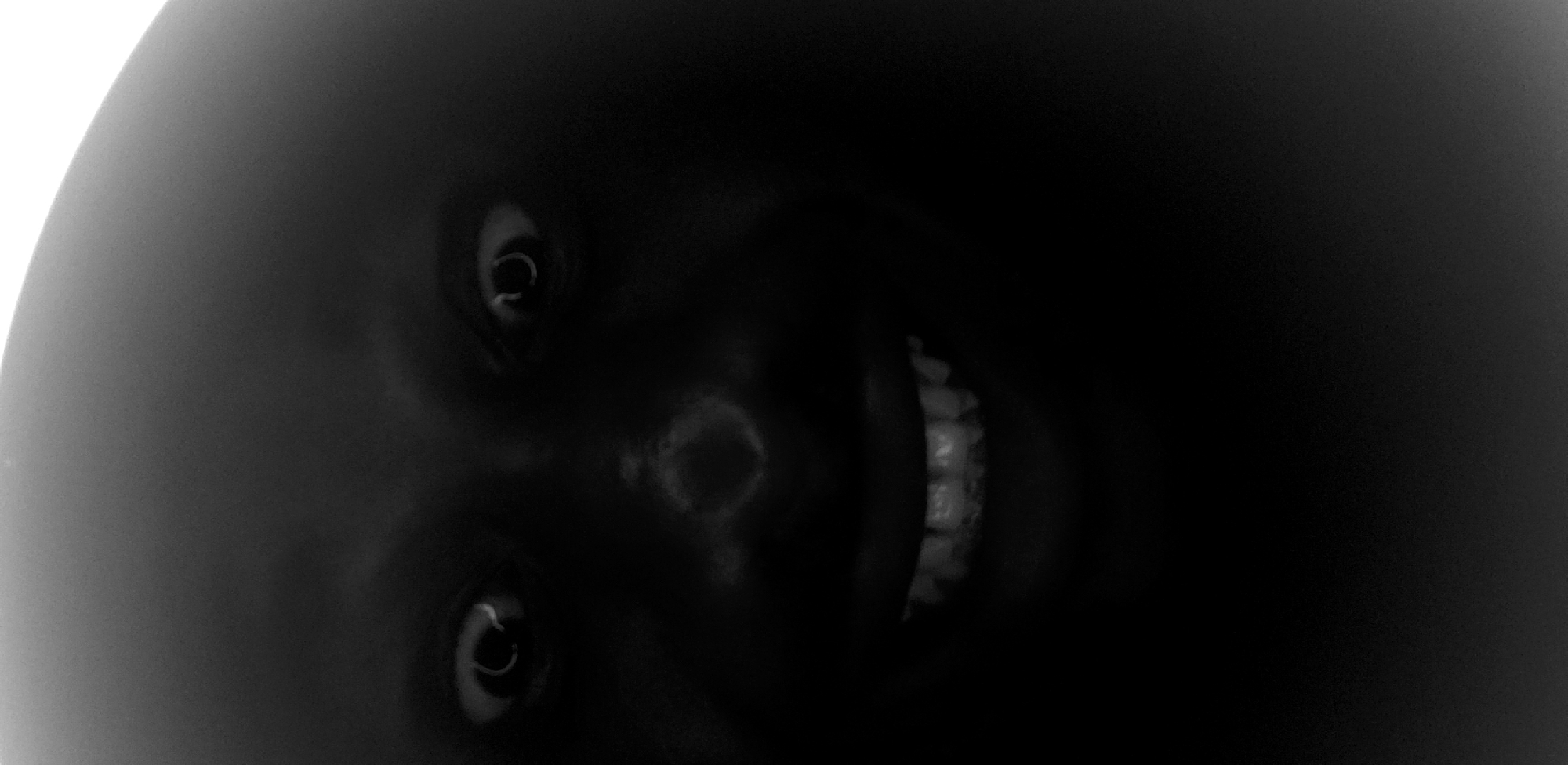 The image shows a close-up, black and white shot of a person's face, predominantly in shadow. Two wide, dark eyes with bright reflections are visible, along with a nose and an open mouth revealing teeth. The overall impression is one of stark contrast and limited detail due to the lighting. There is no discernible indication of the image's location within the city of Narayi, Nigeria, or any environmental context beyond the subject's face.