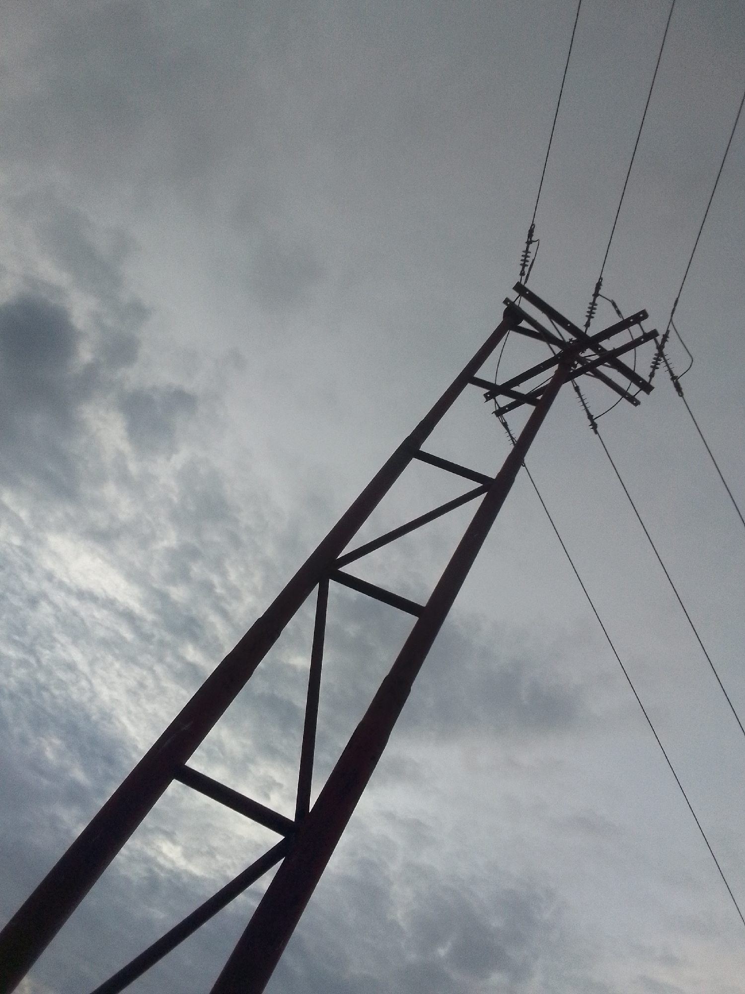 A low-angle shot depicts an electrical transmission tower silhouetted against a cloudy, overcast sky. The tower is constructed of a dark-colored, possibly rusty, metal framework. Three power lines extend from the top of the tower towards the upper right corner of the frame. The insulators connecting the lines to the tower are visible. The sky is filled with gray and white clouds, suggesting potentially inclement weather. The image location is Juan Griego, Venezuela, but there are no other localizing elements within the frame. There are no people or other discernible objects in the scene.