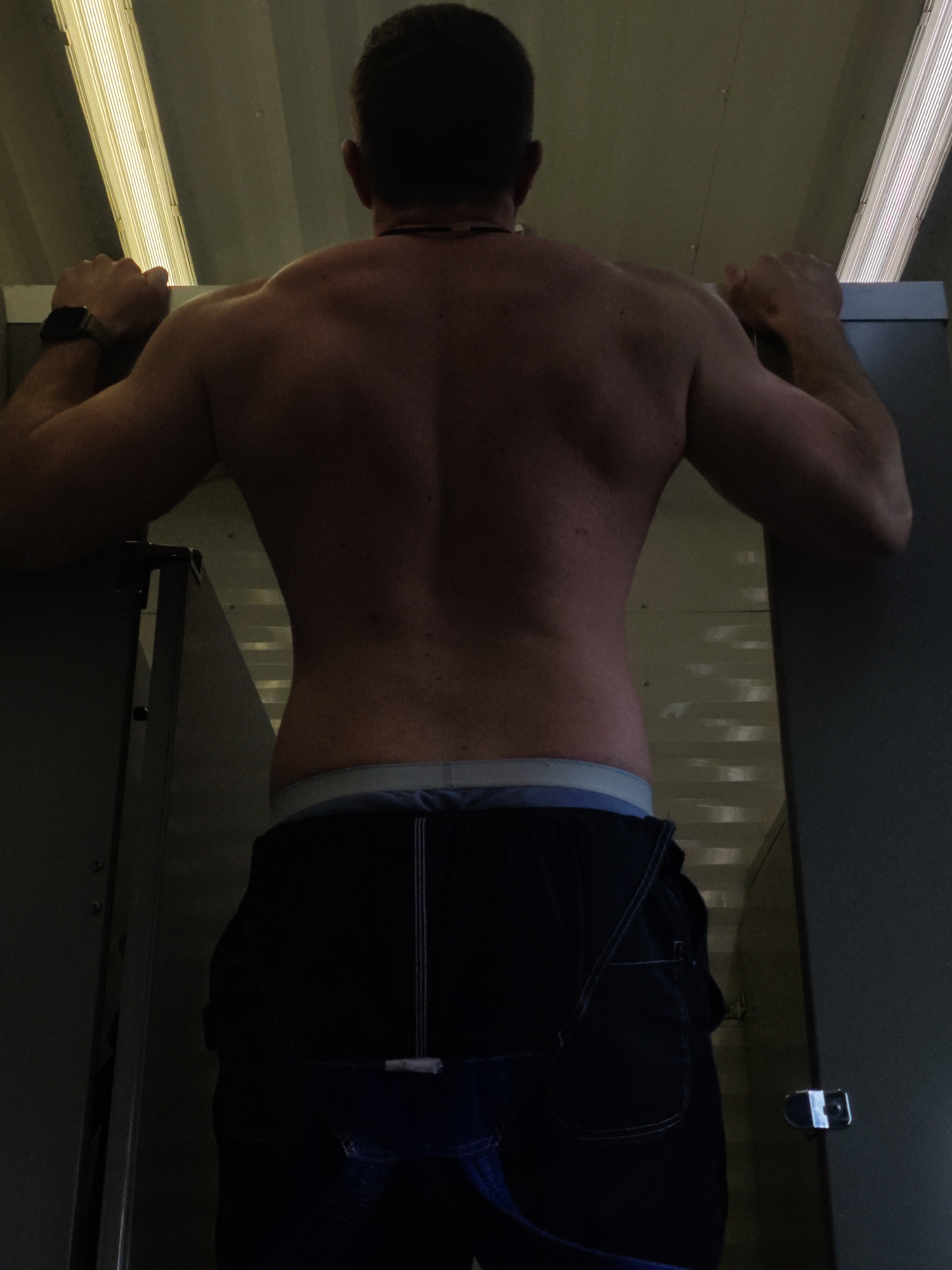 A medium shot captures a muscular man from the back, positioned between two gray, metallic stalls, presumably in a restroom. The man's torso is bare, revealing well-defined back muscles. He wears a black smartwatch on his left wrist and dark-colored pants with visible undergarments. The environment is lit by fluorescent lights overhead, casting a somewhat sterile glow on the scene. The location is likely a public restroom in an urban setting, though the exact location and city are not explicitly defined in the prompt. The absence of additional context or identifying markers prevents a precise geographical location.