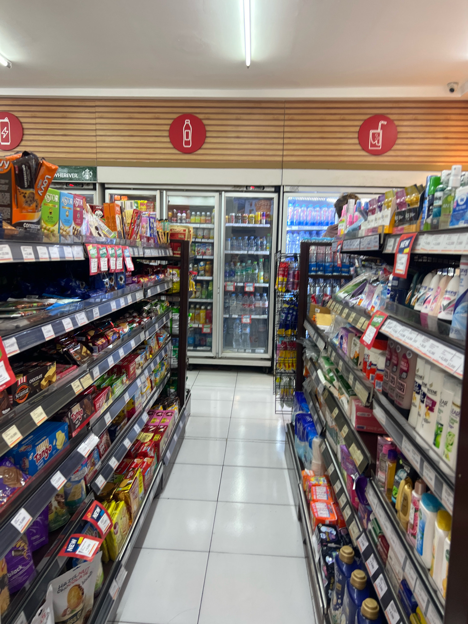 The image shows the interior of a well-lit convenience store, likely a Circle K, with aisles of products on display. The perspective is from an eye-level, wide-angle view looking down a central aisle. On the left, shelves are stocked with various snacks, including chocolates, cookies, and packaged food items. Brands like "Tango" and "Selamat" are visible. In the background, refrigerators display drinks, and promotional signs are present. On the right side, the aisle is lined with shelves containing personal care products such as soaps, shampoos, and lotions, as well as cleaning supplies and detergents. The store's ceiling has fluorescent lighting and what appears to be a security camera. Above the refrigerators, there are horizontal wooden slat walls with circular red icons: one depicting a lightning bolt, another a water bottle, and a third a drinking glass with a straw. The floor is tiled with white square tiles. The environment suggests a typical urban retail space in Denpasar, Indonesia. There are no people clearly visible in the frame, only a blurry figure in the far background behind the refrigerators.