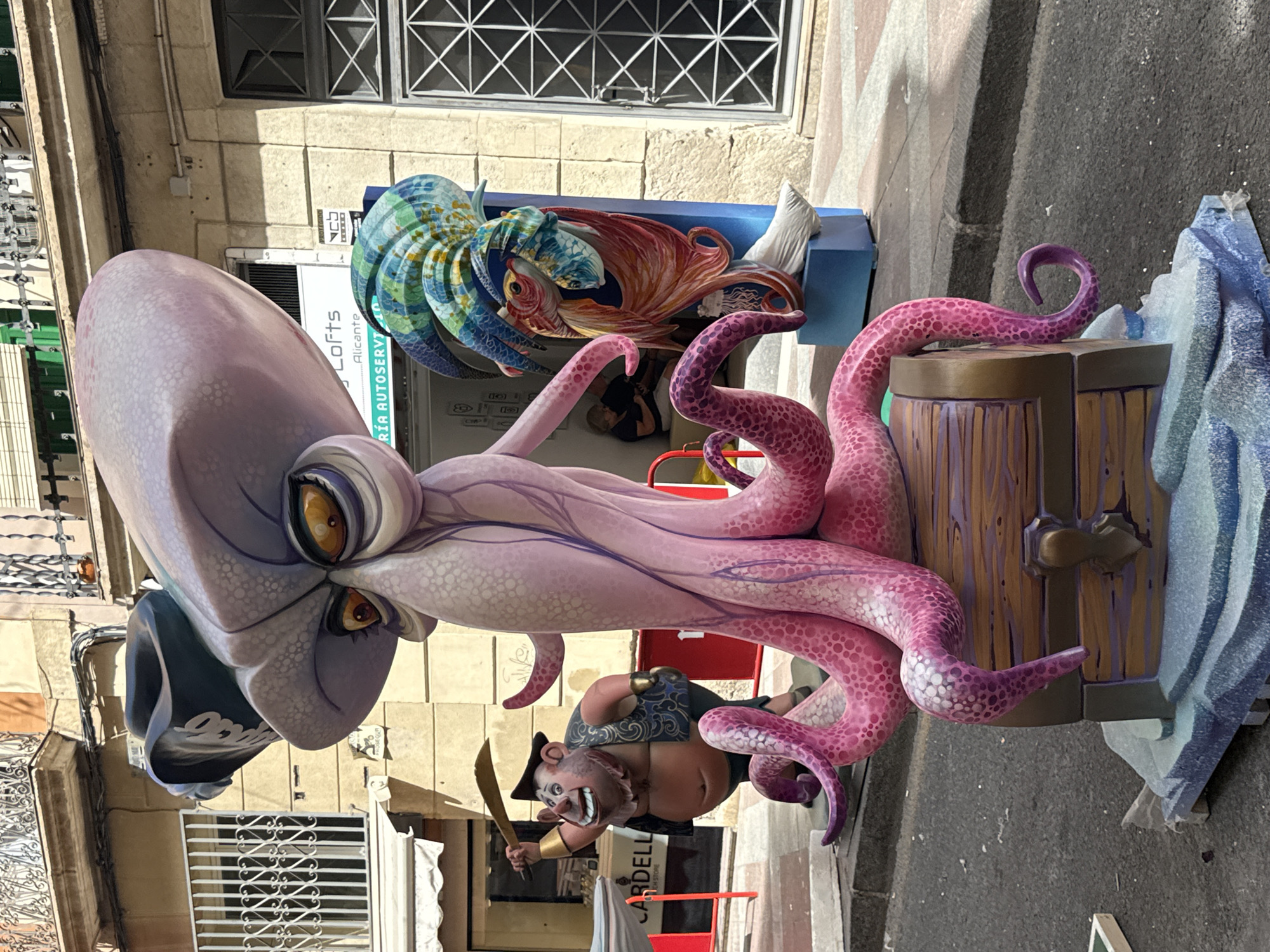 The image depicts an outdoor street scene in front of a building with balconies and windows. The primary subject is a large, whimsical sculpture of a pink octopus wearing a pirate hat, seated on a treasure chest. The octopus has large, expressive yellow eyes and numerous tentacles. To the left of the octopus, there is a smaller sculpture of a caricature-like character, possibly a pirate or sea creature, holding a sword. In the background, another sculpture features colorful, stylized fish and a chameleon. The scene appears to be part of a street festival or exhibition, with the sculptures prominently displayed on the sidewalk and street. The overall environment suggests an urban setting with a festive atmosphere.