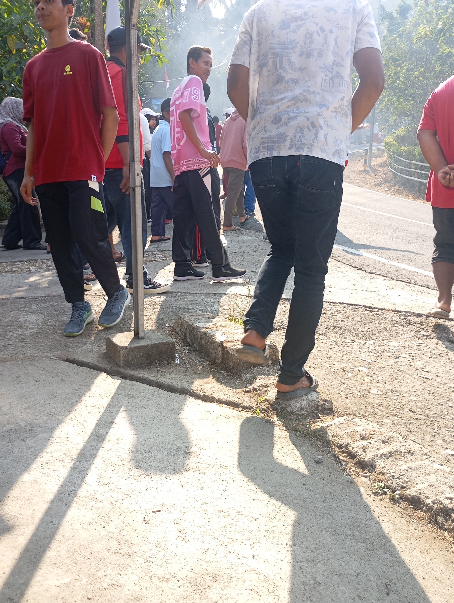 The image captures a sunny daytime scene on a paved pathway next to a road in Ketepung, Indonesia. Several people are present, mostly in the mid-ground and background, walking or standing. In the foreground, a young man wearing a red t-shirt and black pants with a green stripe is standing, casting a shadow. To his right, another young man in a white t-shirt with a blue and white patterned print and black jeans is walking away from the camera. Further back, several other individuals are visible, including a person in a pink t-shirt with the number "75" printed on it, and others in various casual clothing. The environment appears to be outdoors, with trees and foliage visible in the background, suggesting a somewhat rural or suburban setting. The pathway is made of concrete and has some unevenness and cracks. Sunlight creates strong shadows on the ground.
