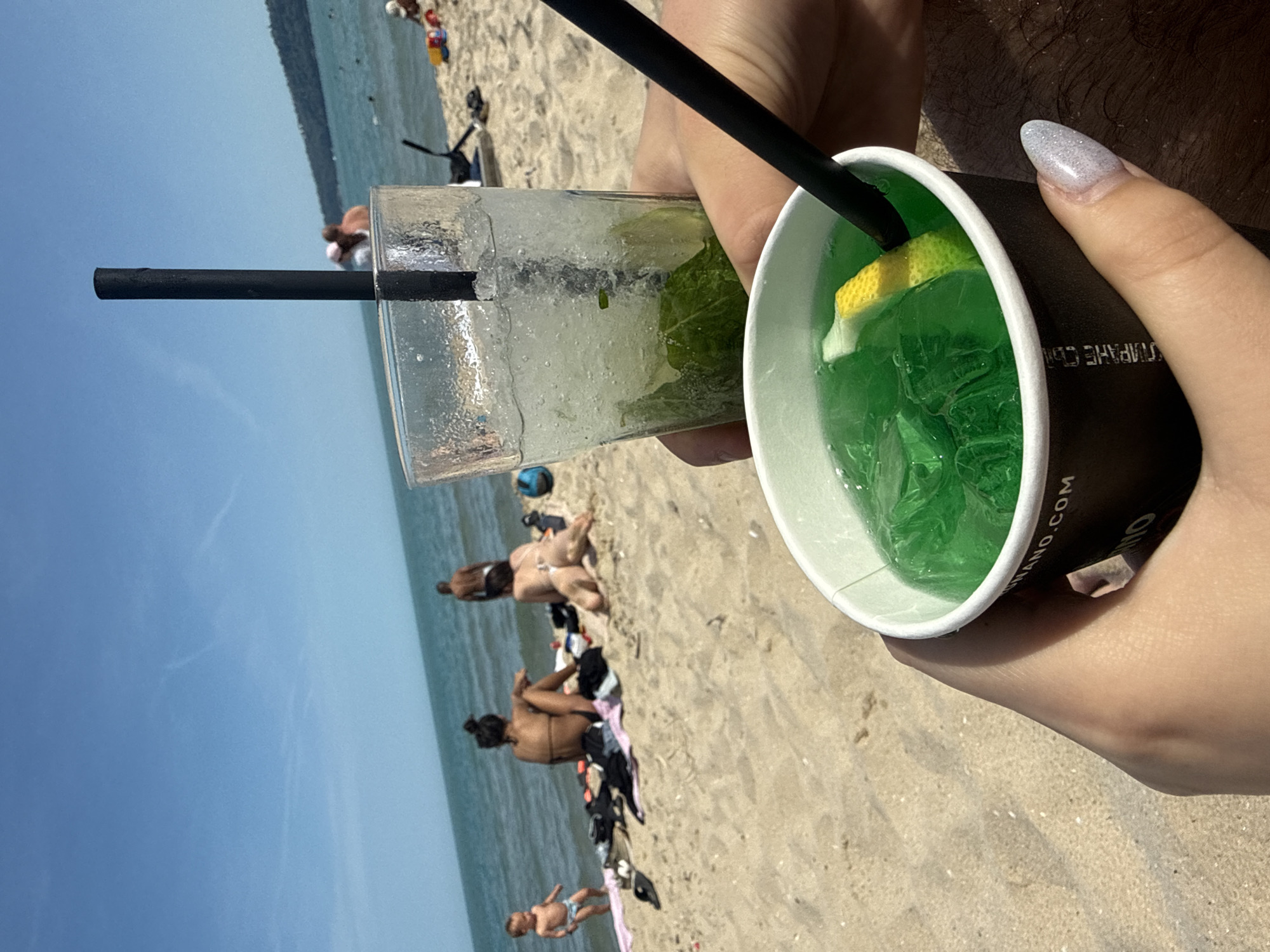 The image depicts a bright, sunny day on a sandy beach. In the immediate foreground, two hands are holding beverages. The left hand holds a black paper cup containing a bright green liquid with ice cubes and a lemon wedge, with a black straw inserted. Partial text "ANO.COM" and Cyrillic characters are visible on the cup. The right hand, adorned with glittery white, almond-shaped fingernails, holds a clear glass filled with crushed ice, mint leaves, and a clear or pale green liquid, also with a black straw.
Behind the hands and drinks, the midground reveals a beach scene. The light-colored sand extends towards the water. On the sand, a woman with dark hair tied up is seated, facing the sea, wearing a dark bikini top. Nearby, another woman is lying prone on her stomach, facing away from the camera towards the water, wearing light-colored bikini bottoms. To the far left, a young child in a light-colored diaper or swim bottom stands at the water's edge, facing left. Various personal items, including clothing and towels, are scattered on the sand near the seated woman. Further right, a shirtless man stands in the shallow water, facing away from the camera.
The background features a calm, light blue-green sea under a clear, bright blue sky with faint cirrus clouds or contrails. In the far distance on the right, a dark green landmass, possibly hills or a forested area, is visible along the coastline. The scene is illuminated by direct sunlight, characteristic of midday. The location is a sandy beach next to a sea or ocean, in the city of Unknown, Unknown.