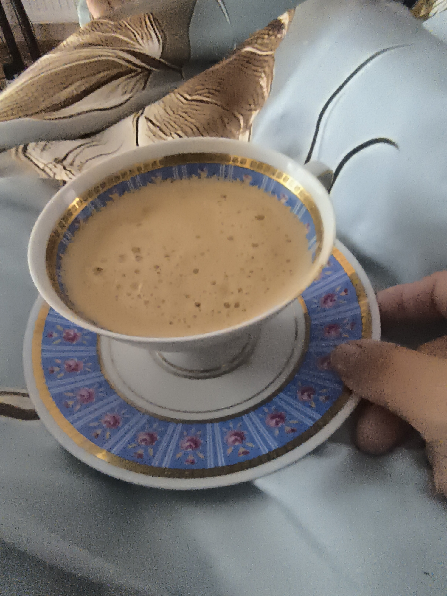 A close-up, slightly blurry, low-angle shot shows a person's hand holding a teacup and saucer on a pale blue, textured surface, possibly a bed. The teacup is filled with a frothy, beige liquid, likely coffee or tea, and sits on a saucer with a decorative blue band featuring pink floral designs and gold trim. A glimpse of what appears to be a patterned blanket or fabric is visible in the upper left quadrant of the image. The lighting suggests an indoor setting, potentially a bedroom or living area. There is no indication of the specific location being Klaipėda, Lithuania.