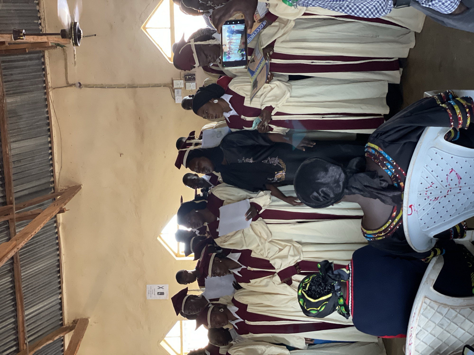 This media file shows a group of people, likely a choir, dressed in matching robes with maroon and cream accents. The robes are long and appear to be ceremonial or formal. The individuals are standing or sitting in what seems to be an indoor setting, possibly a church or community hall, given the simple construction with exposed wooden beams and corrugated metal roofing visible in some parts of the image. A ceiling fan is mounted on the wooden beams.
The lighting suggests it could be daytime, with natural light entering from windows. One person in the foreground is holding up a mobile phone to take a picture or video of the group. Some individuals are holding papers, perhaps song sheets or hymnals. A small sign with an 'X' and some text is visible on the wall, which might indicate a prohibition of some activity, but the text is not legible. The expressions on the faces of the people are generally neutral, and the overall atmosphere appears calm and orderly. The scene is consistent with a choir practice, a performance, or a religious gathering in Kassa, Nigeria.