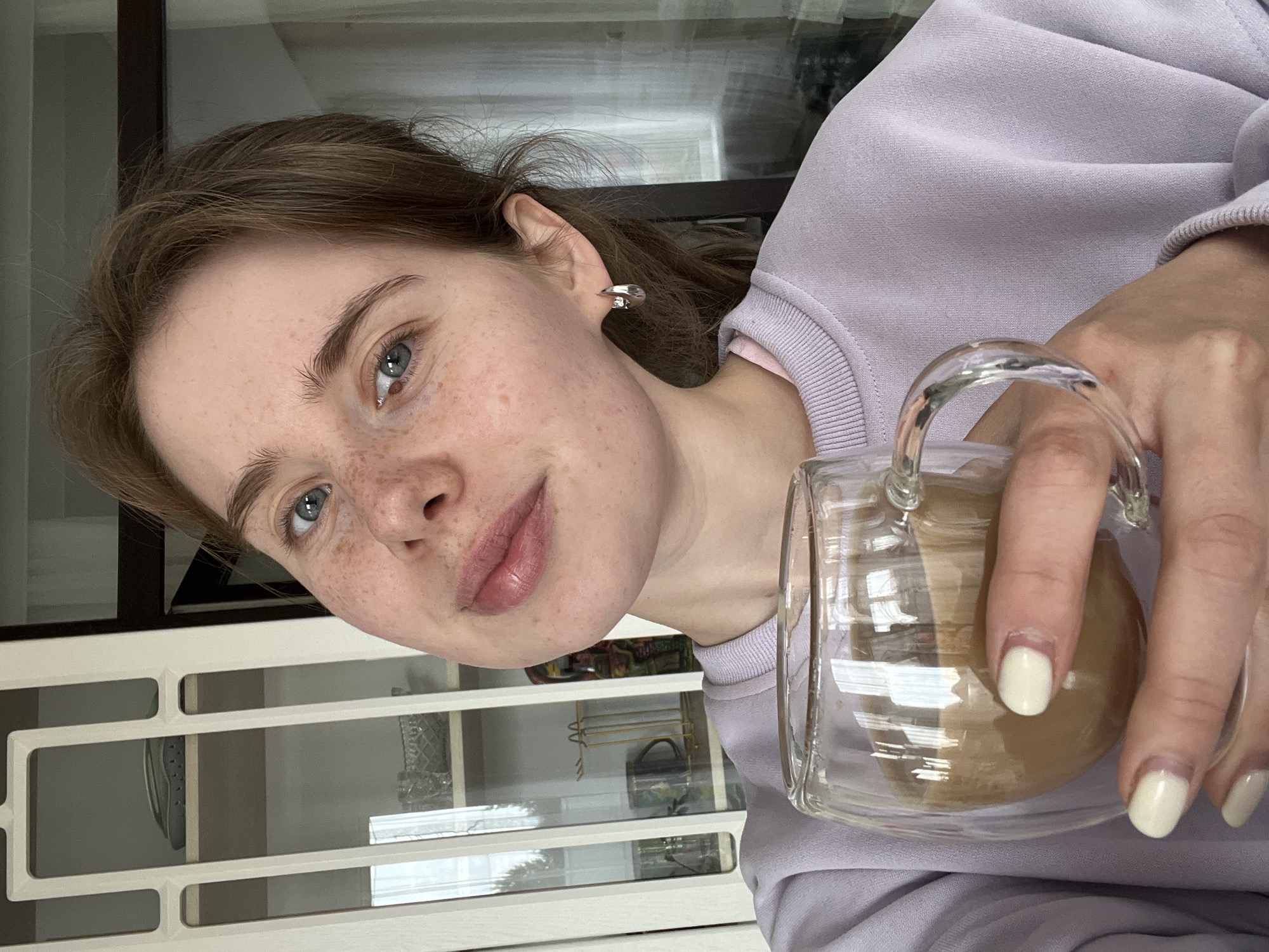 A young woman with fair skin, light brown hair, and freckles is holding a glass mug with a light-colored beverage inside, likely coffee or tea. She is wearing a lilac-colored sweatshirt. Her fingernails are painted a pale cream color. A silver earring is visible in her left ear.
Behind her is a white cabinet or shelving unit with glass doors. The reflection in the glass doors suggests that the setting might be indoors, possibly near a window. It is not possible to confirm the location is in Chaikovsky, Russia, from the information in the image.