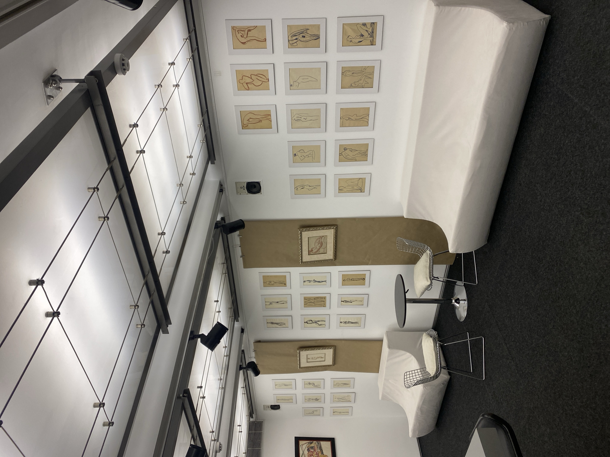 The image shows the interior of what appears to be a gallery or showroom. The location is unknown.
The upper section of the frame shows a white wall with a series of framed sketches of human figures. A long, white bench-like structure is placed along the wall.
The lower section mirrors the upper section with another set of framed sketches on the wall, interspersed with strips of tan-colored material. A small table with two chairs are positioned near the wall, and a larger framed picture is visible towards the bottom center. A section of what appears to be a dark carpet covers the floor.
The image appears to be taken from an elevated position looking downwards.