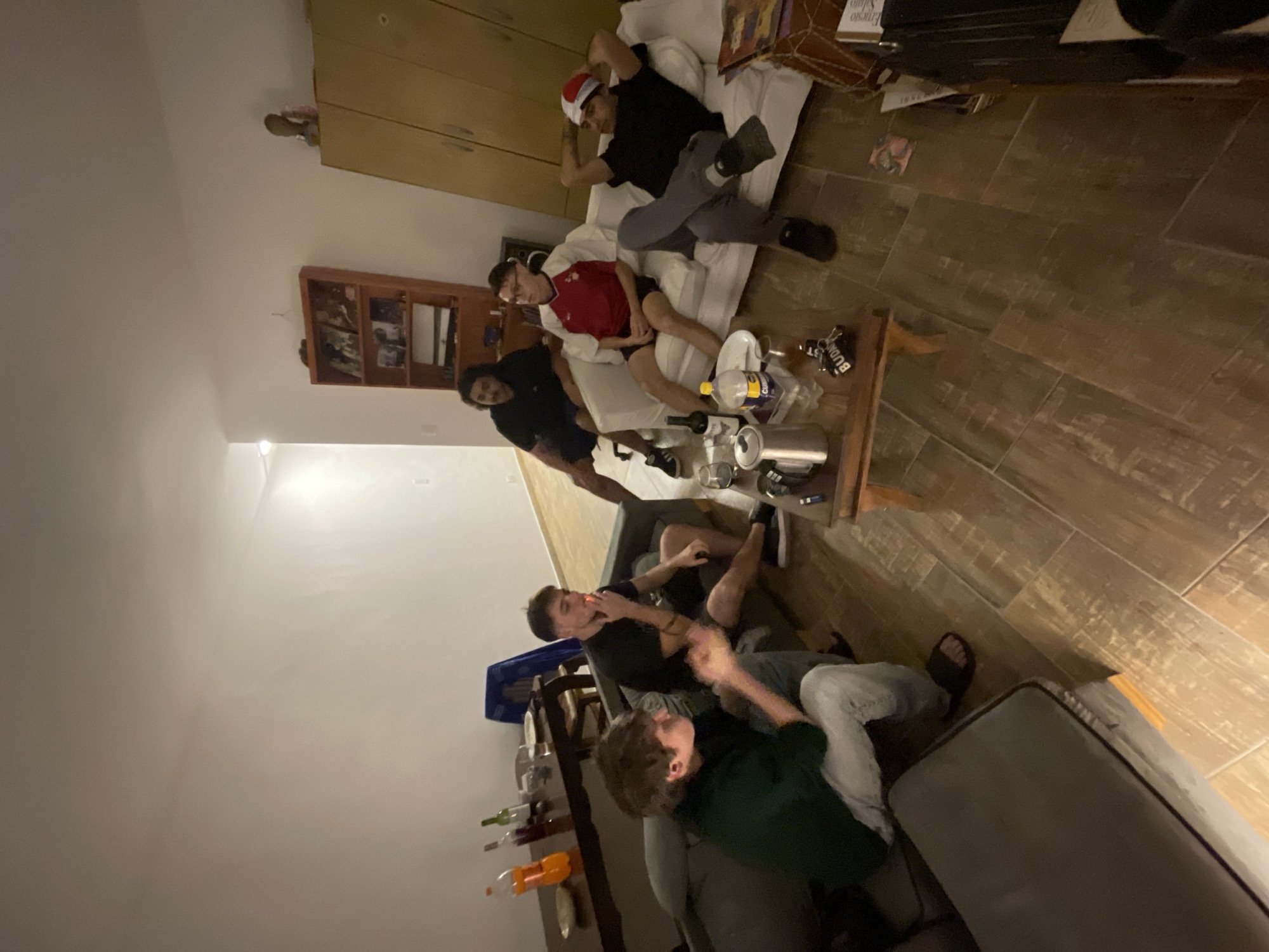 An informal gathering of at least six young men is depicted in an indoor setting, likely a living room, at night or in the evening. The photograph is taken from a high angle and rotated, suggesting a casual, spontaneous moment in Buenos Aires, Argentina.
The scene features two sofas. On a light-colored sofa, one man in a black shirt and grey pants is lying down, wearing a red and white Santa hat. Next to him, another man in a red shirt and glasses is seated, and a third man in a black shirt is also on the sofa. Further into the room, on a darker grey sofa, two more men are visible. One, wearing a black t-shirt, is seen smoking, while the other, in a dark green long-sleeved shirt and light pants, sits beside him.
A wooden coffee table in the foreground holds a large plastic bottle (with "CLight" visible on its label), several aluminum cans, and a glass bottle. Another wooden table in the mid-ground also holds bottles, including one with orange liquid, and plates. The floor is covered with wood-look tiles. A black slipper and a keychain with the partially visible text "BUDG T" are on the floor. In the background, a wooden cabinet/shelf unit is visible, holding various items. The atmosphere appears relaxed and social, consistent with friends hanging out, possibly during the Christmas season given the Santa hat.